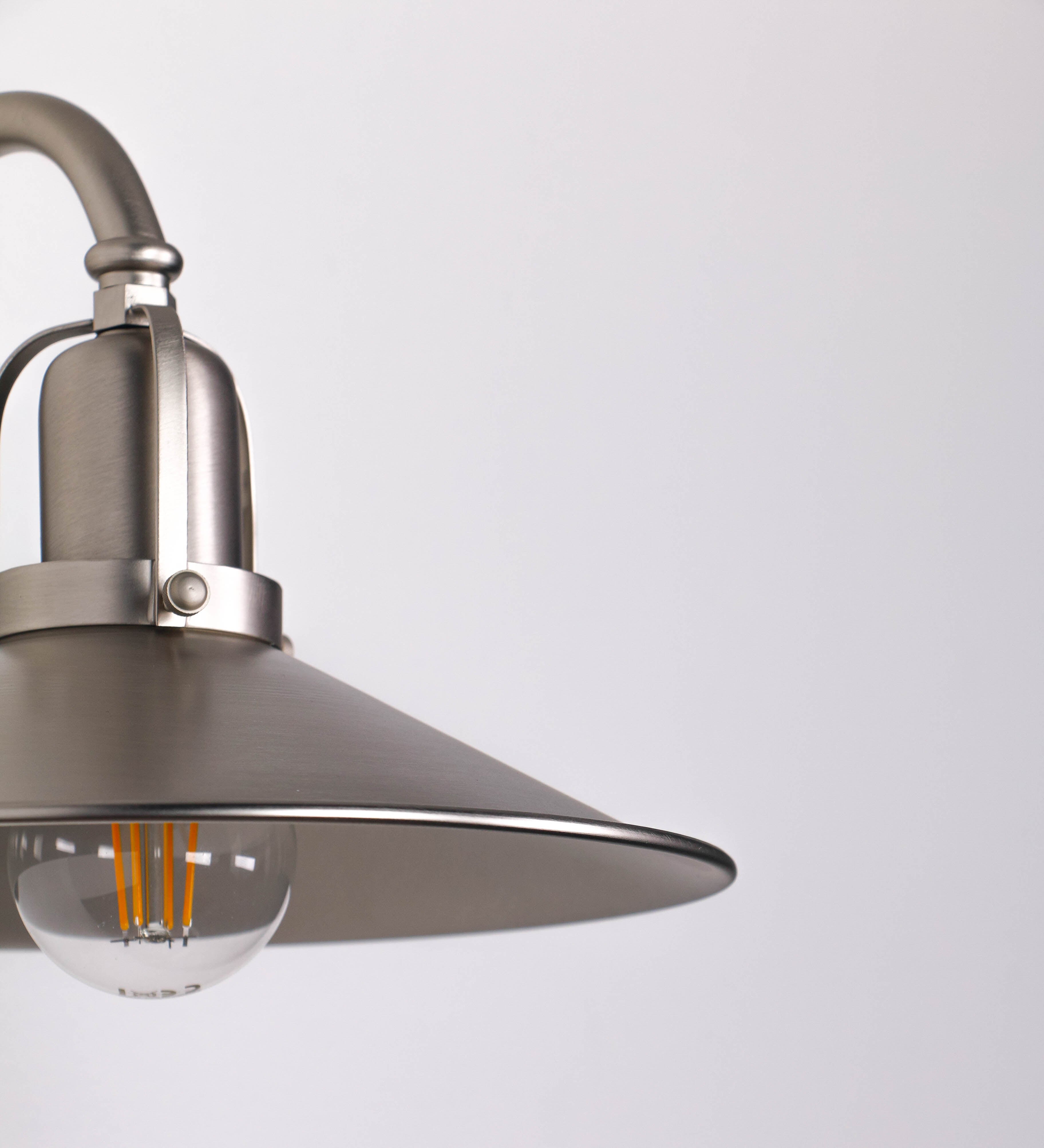 Aplique de parede todo em níquel escovado, com um design clássico e cores contemporâneas. O aplique é composto por uma base circular, de onde se estende um braço curvo, que forma um arco gracioso e sustenta o chapéu do candeeiro. O chapéu tem um formato cónico invertido, com uma borda ligeiramente levantada, que direciona a luz para baixo. No topo do chapéu, há um suporte circular com três braços que fixa o chapéu ao braço curvo.