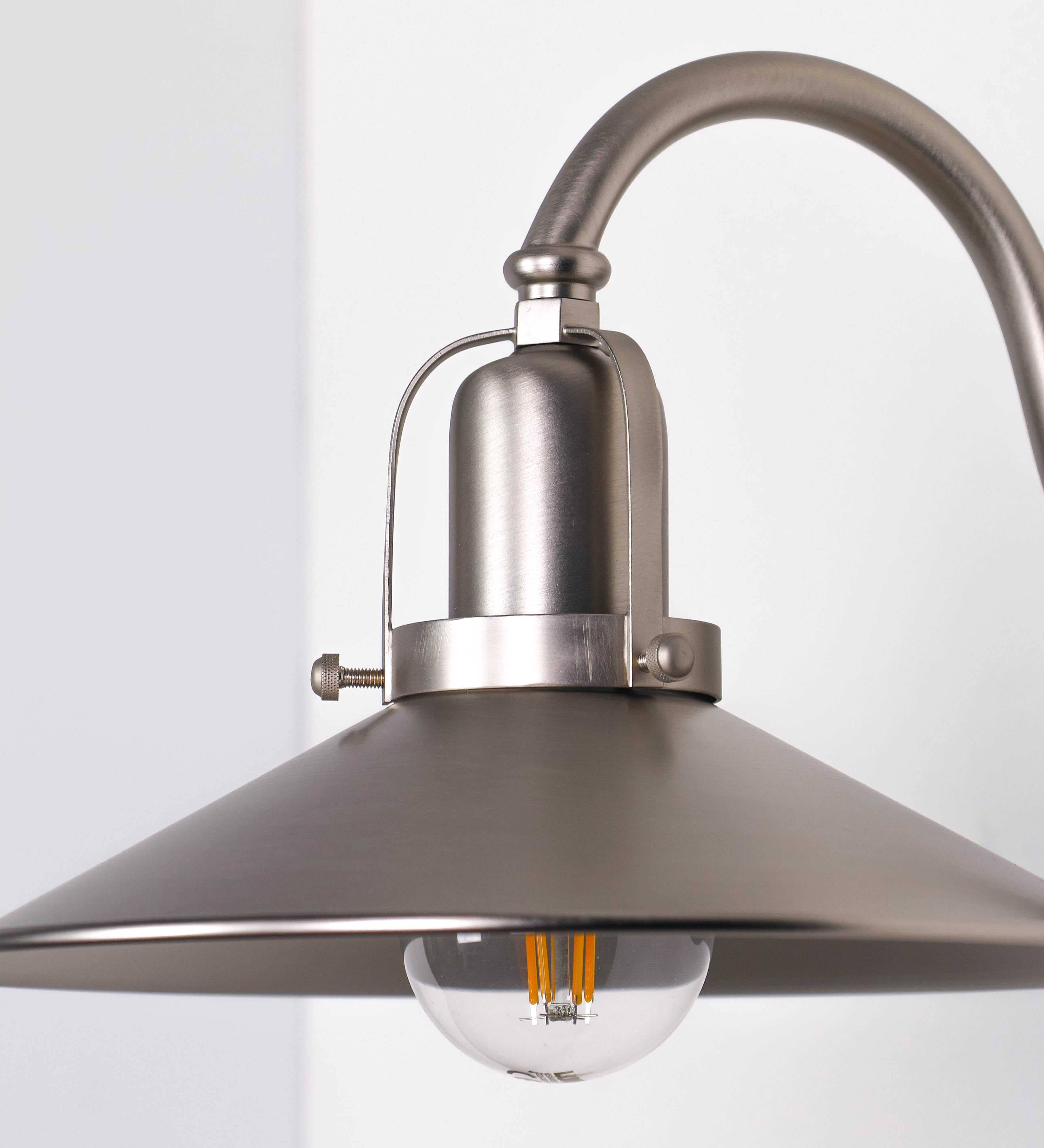 Aplique de parede todo em níquel escovado, com um design clássico e cores contemporâneas. O aplique é composto por uma base circular, de onde se estende um braço curvo, que forma um arco gracioso e sustenta o chapéu do candeeiro. O chapéu tem um formato cónico invertido, com uma borda ligeiramente levantada, que direciona a luz para baixo. No topo do chapéu, há um suporte circular com três braços que fixa o chapéu ao braço curvo.