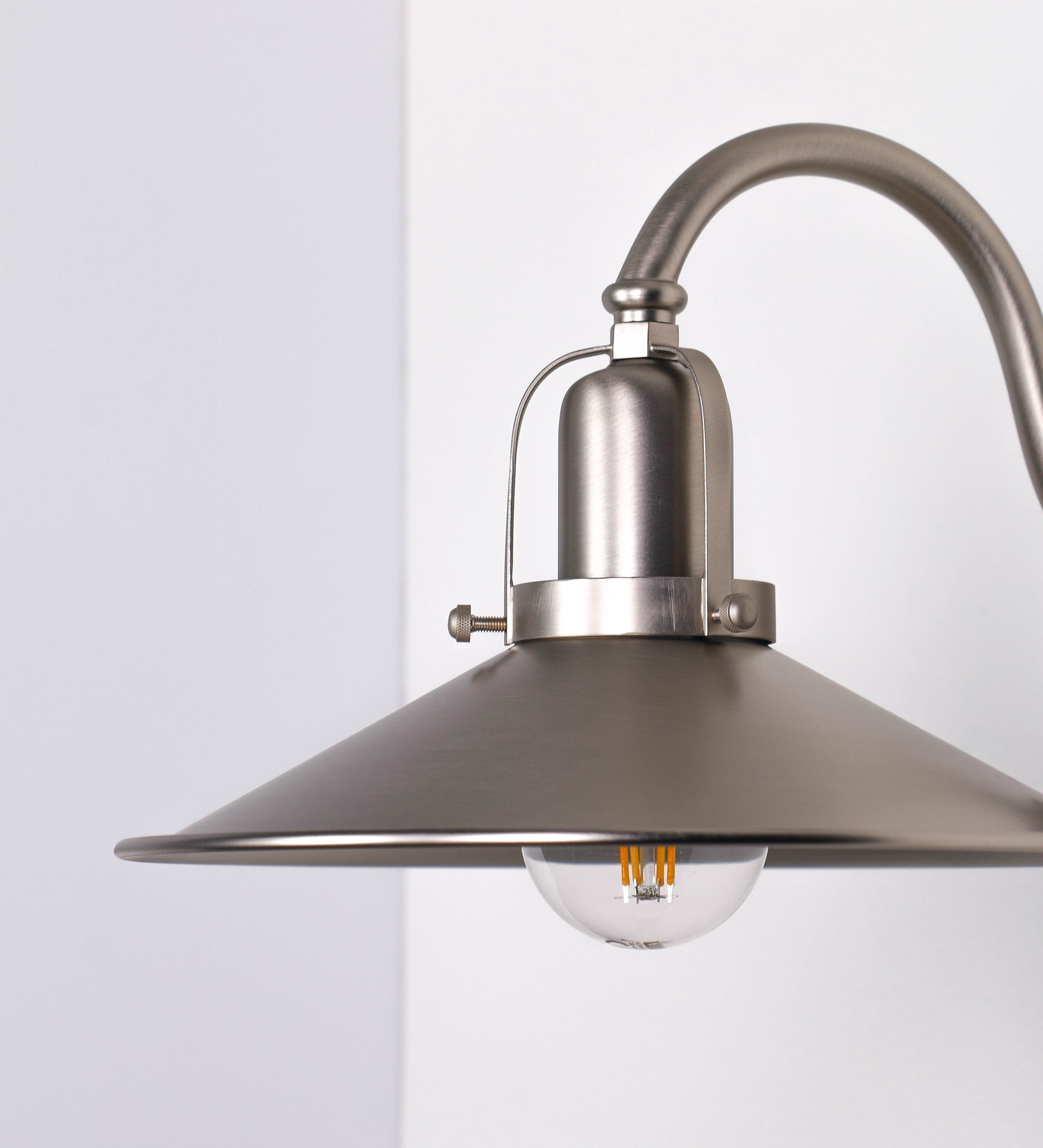 Aplique de parede todo em níquel escovado, com um design clássico e cores contemporâneas. O aplique é composto por uma base circular, de onde se estende um braço curvo, que forma um arco gracioso e sustenta o chapéu do candeeiro. O chapéu tem um formato cónico invertido, com uma borda ligeiramente levantada, que direciona a luz para baixo. No topo do chapéu, há um suporte circular com três braços que fixa o chapéu ao braço curvo.