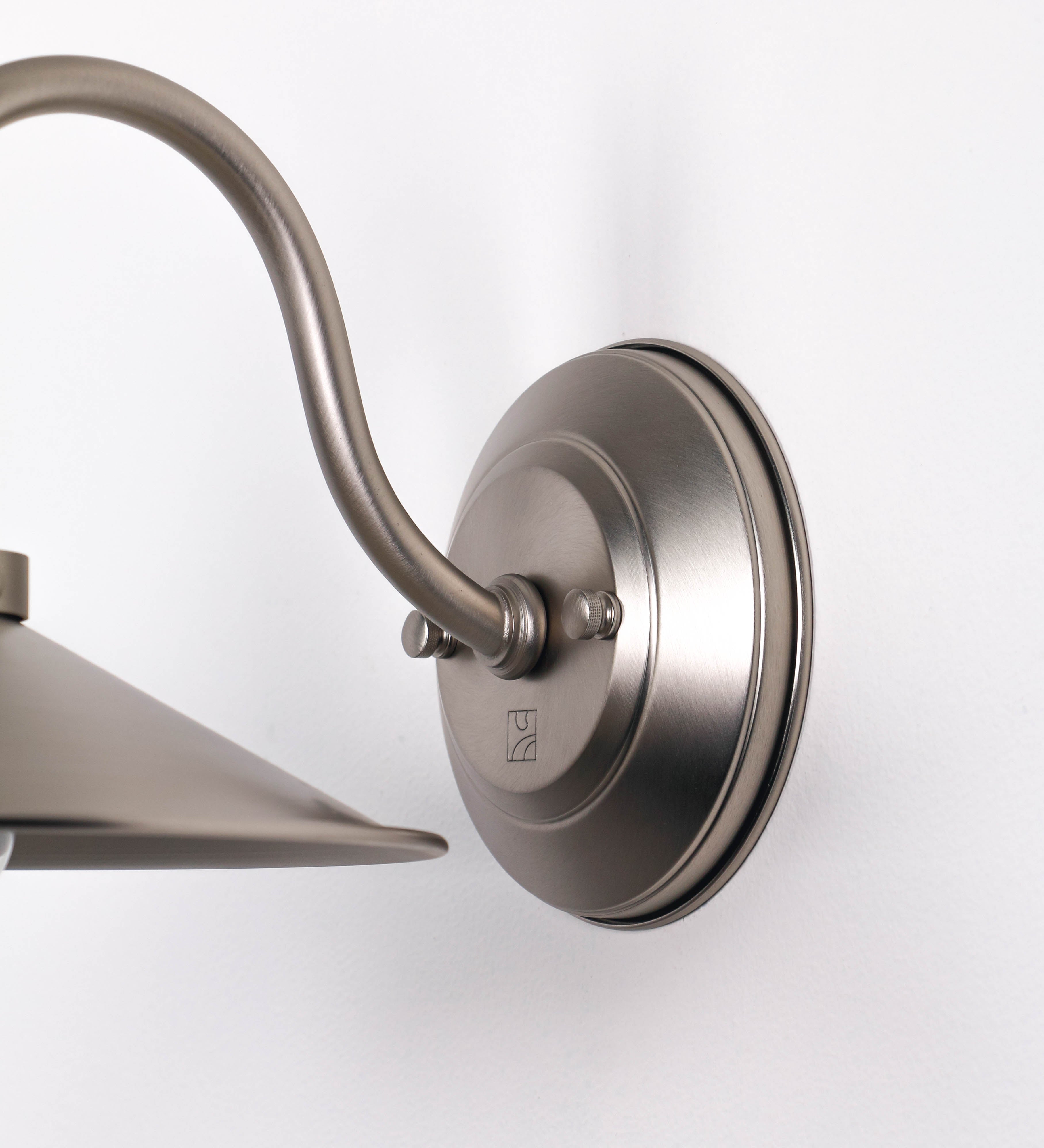 Aplique de parede todo em níquel escovado, com um design clássico e cores contemporâneas. O aplique é composto por uma base circular, de onde se estende um braço curvo, que forma um arco gracioso e sustenta o chapéu do candeeiro. O chapéu tem um formato cónico invertido, com uma borda ligeiramente levantada, que direciona a luz para baixo. No topo do chapéu, há um suporte circular com três braços que fixa o chapéu ao braço curvo.