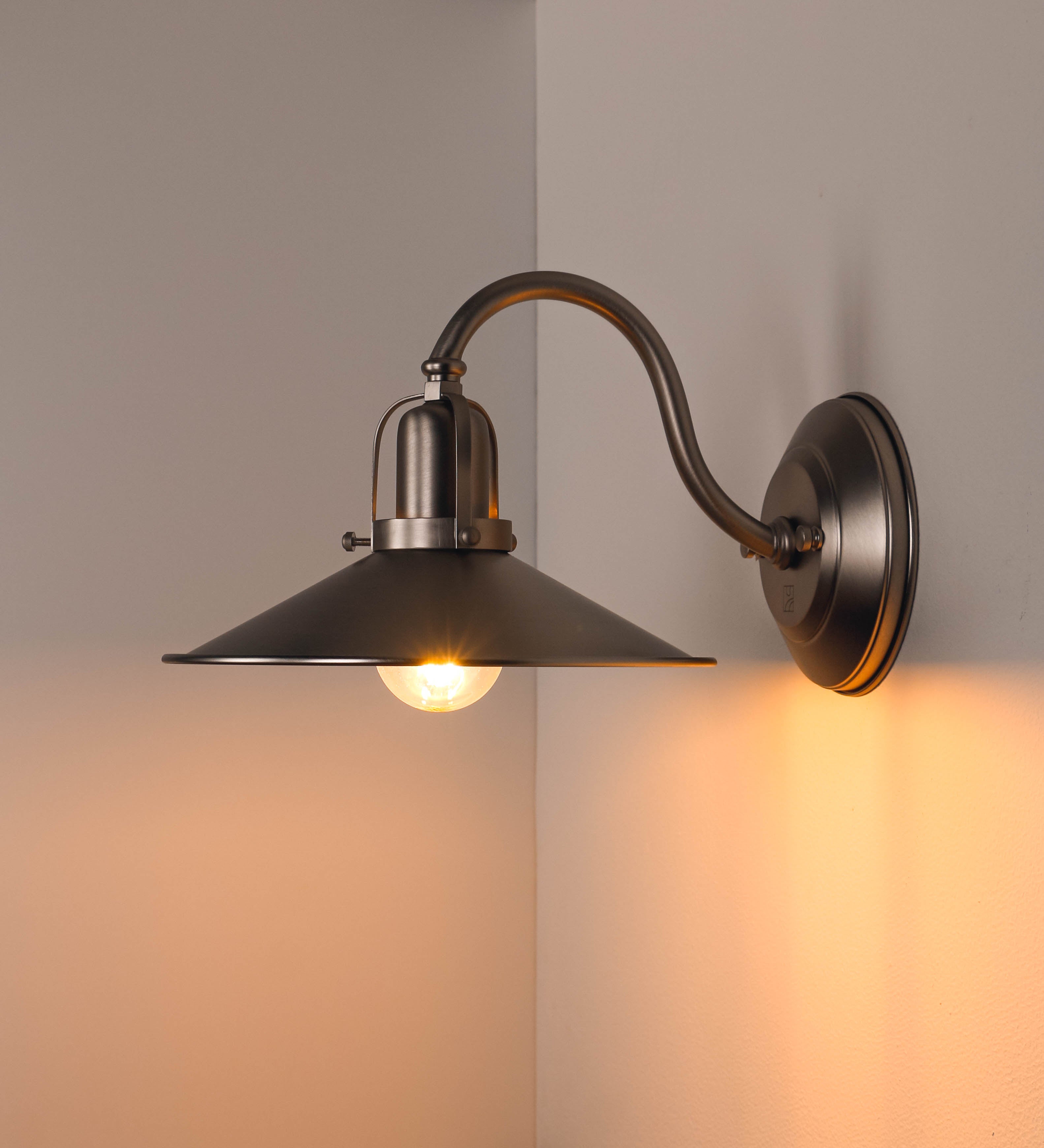 Aplique de parede todo em níquel escovado, com um design clássico e cores contemporâneas. O aplique é composto por uma base circular, de onde se estende um braço curvo, que forma um arco gracioso e sustenta o chapéu do candeeiro. O chapéu tem um formato cónico invertido, com uma borda ligeiramente levantada, que direciona a luz para baixo. No topo do chapéu, há um suporte circular com três braços que fixa o chapéu ao braço curvo.