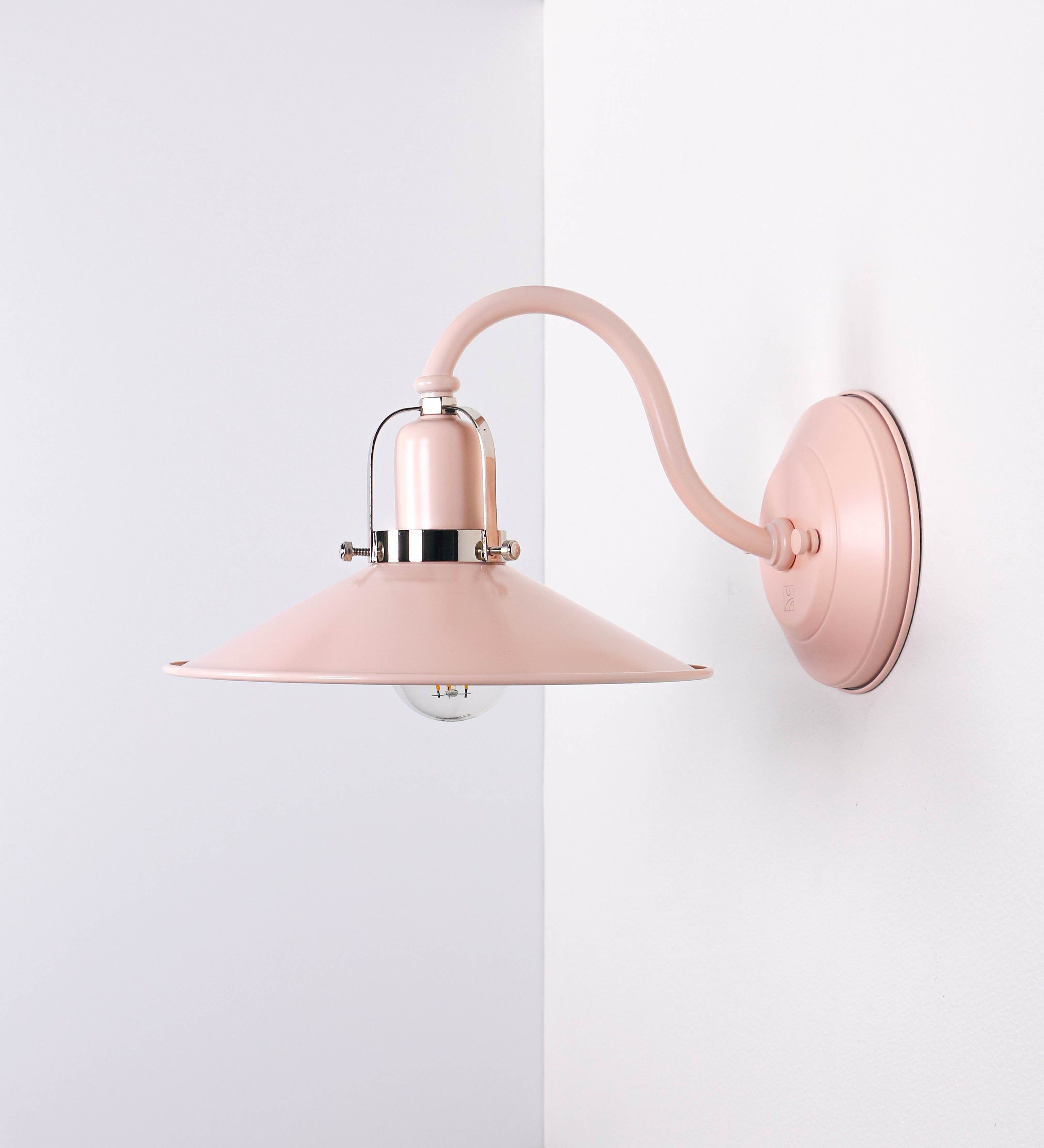 Aplique de parede com um design clássico e cores contemporâneas. O aplique é composto por uma base circular rosa clara, de onde se estende um braço curvo, que forma um arco gracioso e sustenta o chapéu do candeeiro, rosa claro também. O chapéu tem um formato cónico invertido, com uma borda ligeiramente levantada, que direciona a luz para baixo. O interior do chapéu é branco. No topo do chapéu, há um suporte circular em níquel polido, com três braços que fixa o chapéu ao braço curvo.