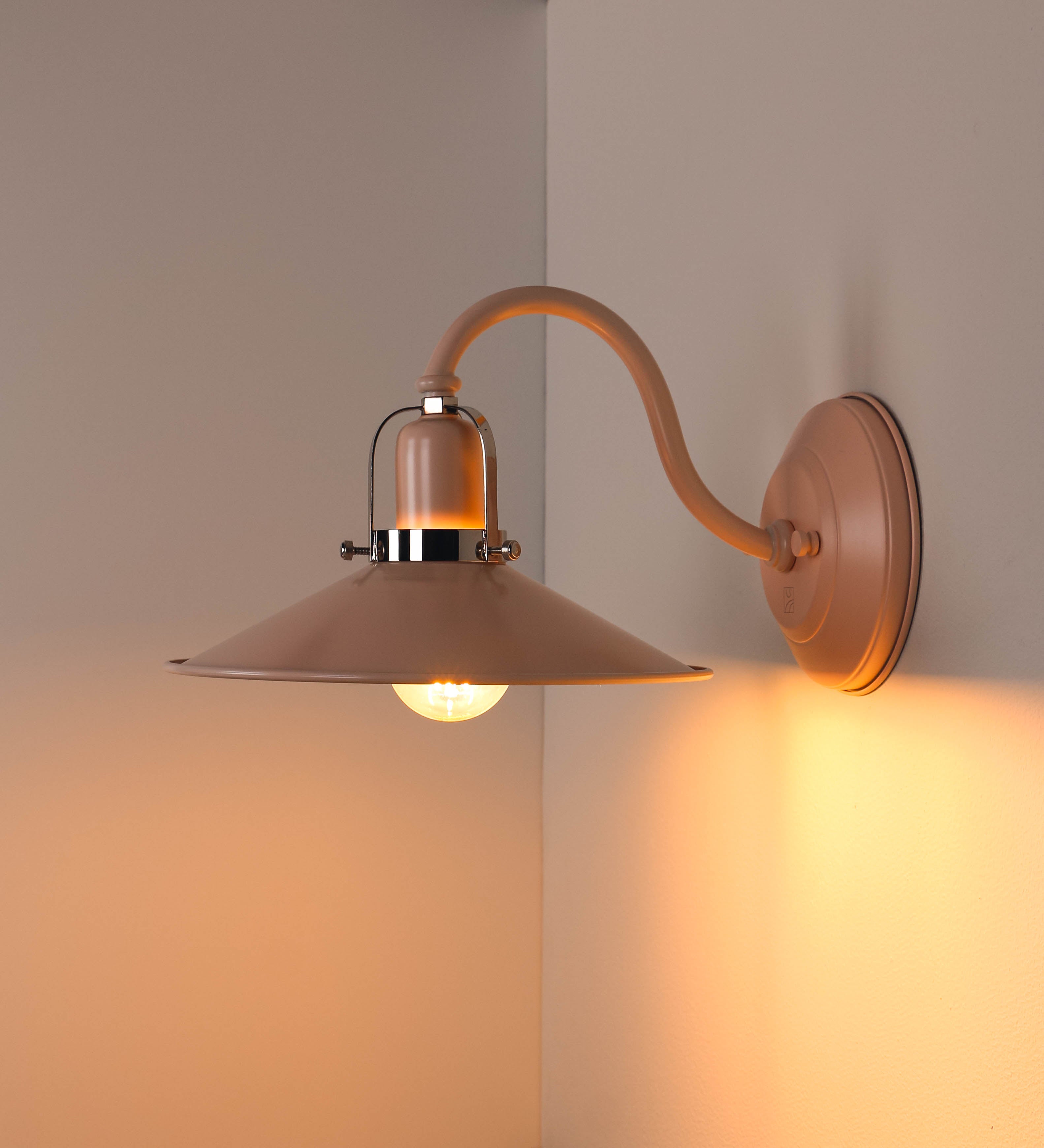 Aplique de parede com um design clássico e cores contemporâneas. O aplique é composto por uma base circular rosa clara, de onde se estende um braço curvo, que forma um arco gracioso e sustenta o chapéu do candeeiro, rosa claro também. O chapéu tem um formato cónico invertido, com uma borda ligeiramente levantada, que direciona a luz para baixo. O interior do chapéu é branco. No topo do chapéu, há um suporte circular em níquel polido, com três braços que fixa o chapéu ao braço curvo.