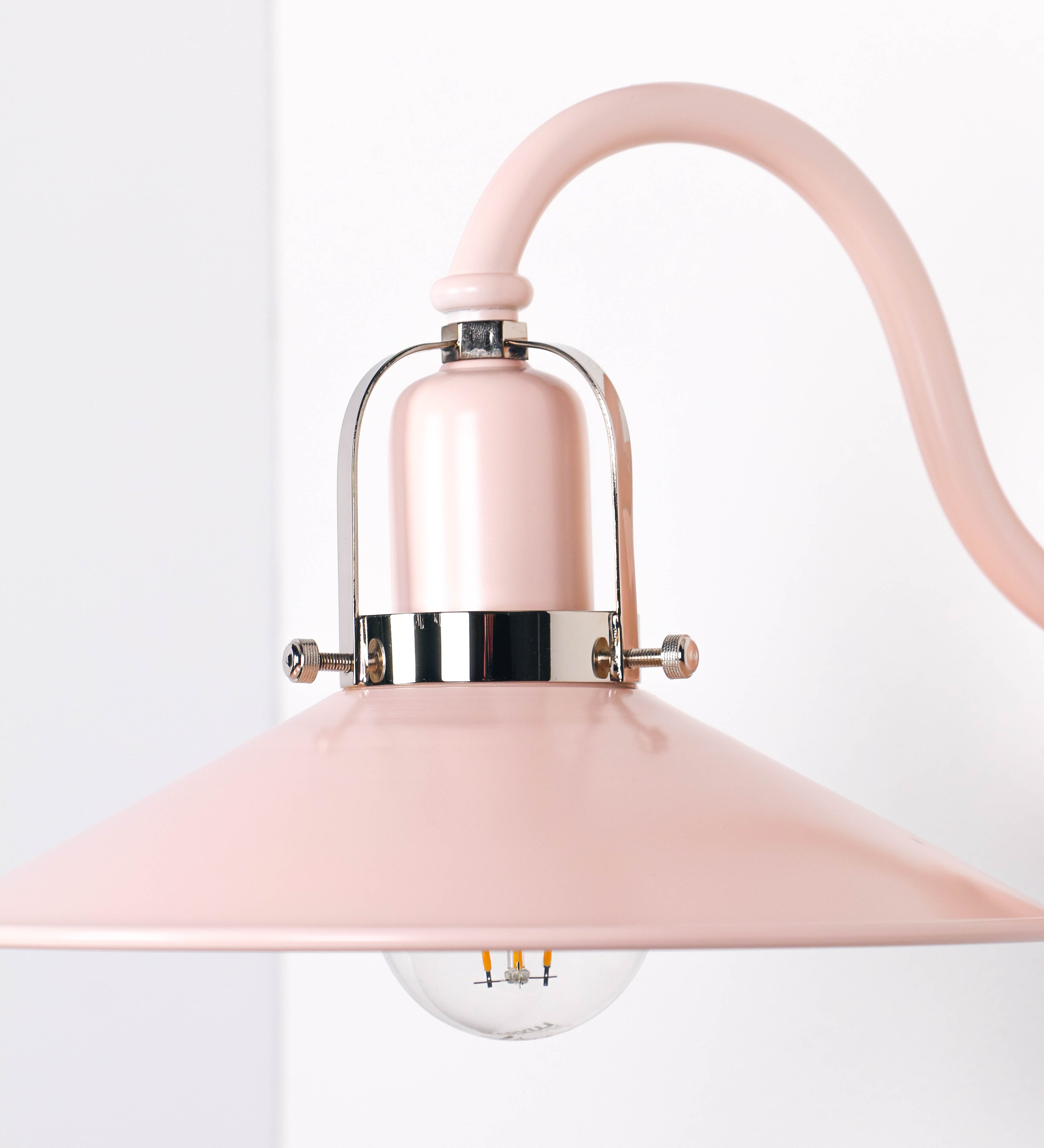 Aplique de parede com um design clássico e cores contemporâneas. O aplique é composto por uma base circular rosa clara, de onde se estende um braço curvo, que forma um arco gracioso e sustenta o chapéu do candeeiro, rosa claro também. O chapéu tem um formato cónico invertido, com uma borda ligeiramente levantada, que direciona a luz para baixo. O interior do chapéu é branco. No topo do chapéu, há um suporte circular em níquel polido, com três braços que fixa o chapéu ao braço curvo.