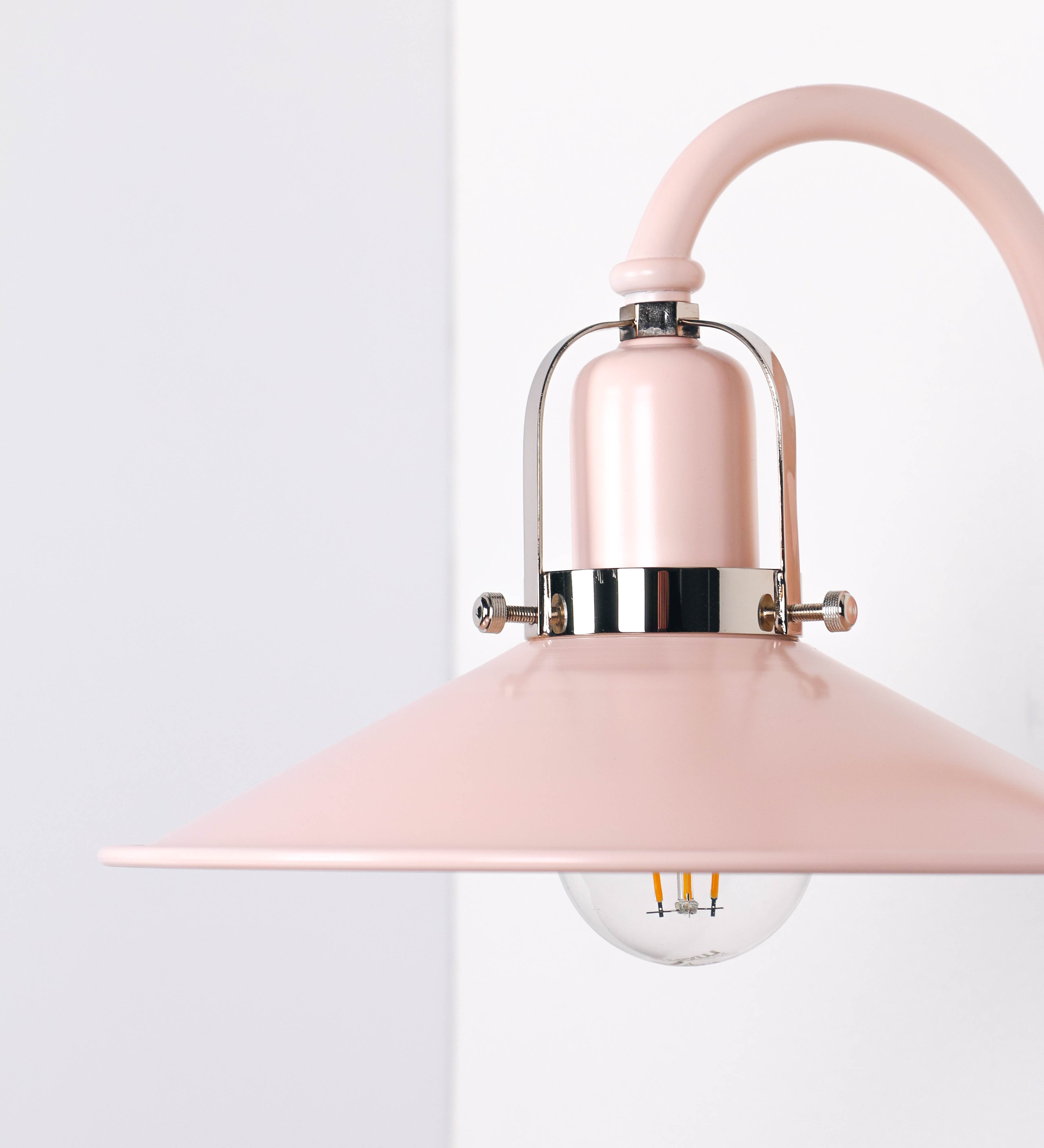 Aplique de parede com um design clássico e cores contemporâneas. O aplique é composto por uma base circular rosa clara, de onde se estende um braço curvo, que forma um arco gracioso e sustenta o chapéu do candeeiro, rosa claro também. O chapéu tem um formato cónico invertido, com uma borda ligeiramente levantada, que direciona a luz para baixo. O interior do chapéu é branco. No topo do chapéu, há um suporte circular em níquel polido, com três braços que fixa o chapéu ao braço curvo.