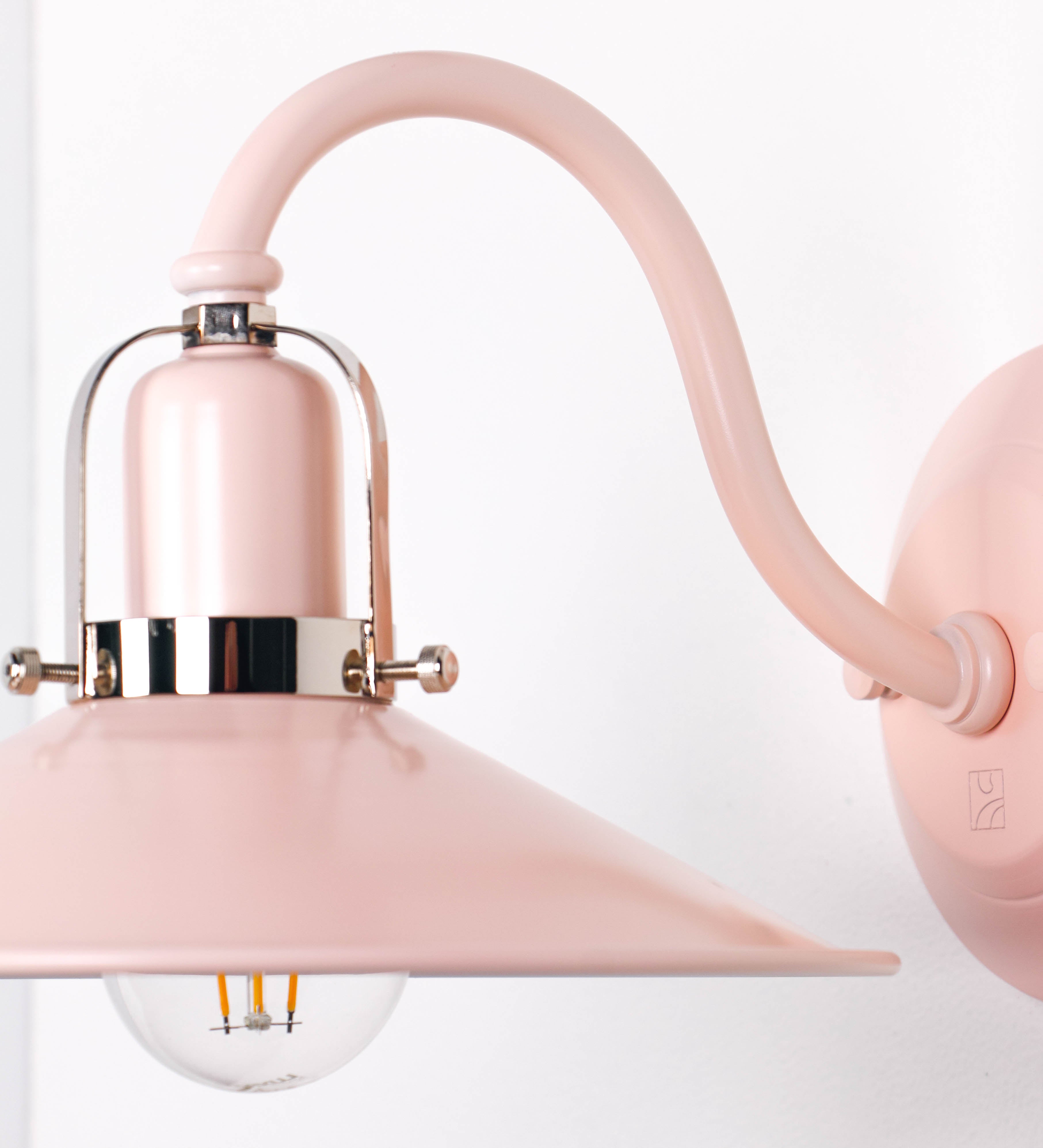 Aplique de parede com um design clássico e cores contemporâneas. O aplique é composto por uma base circular rosa clara, de onde se estende um braço curvo, que forma um arco gracioso e sustenta o chapéu do candeeiro, rosa claro também. O chapéu tem um formato cónico invertido, com uma borda ligeiramente levantada, que direciona a luz para baixo. O interior do chapéu é branco. No topo do chapéu, há um suporte circular em níquel polido, com três braços que fixa o chapéu ao braço curvo.