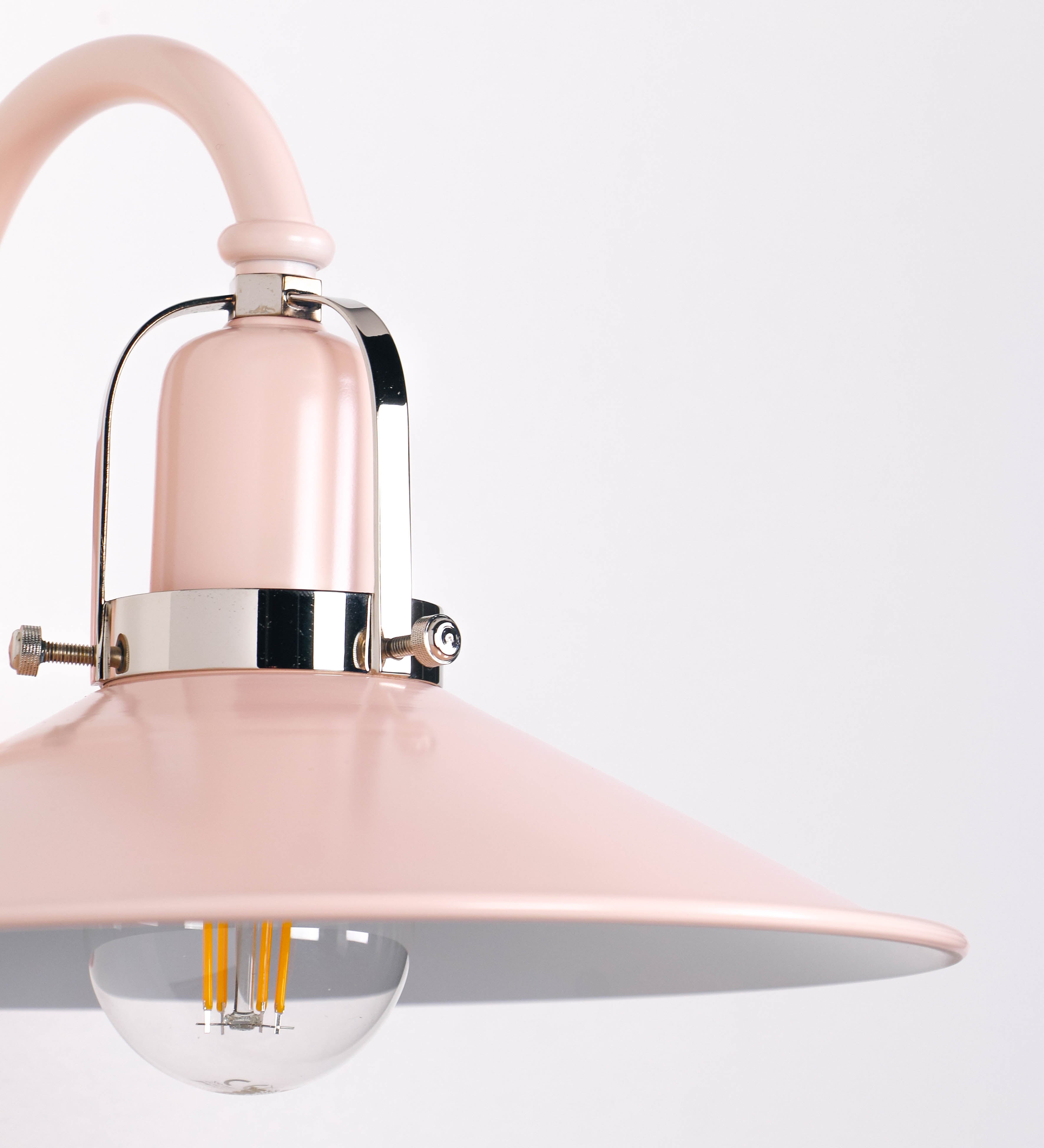 Aplique de parede com um design clássico e cores contemporâneas. O aplique é composto por uma base circular rosa clara, de onde se estende um braço curvo, que forma um arco gracioso e sustenta o chapéu do candeeiro, rosa claro também. O chapéu tem um formato cónico invertido, com uma borda ligeiramente levantada, que direciona a luz para baixo. O interior do chapéu é branco. No topo do chapéu, há um suporte circular em níquel polido, com três braços que fixa o chapéu ao braço curvo.