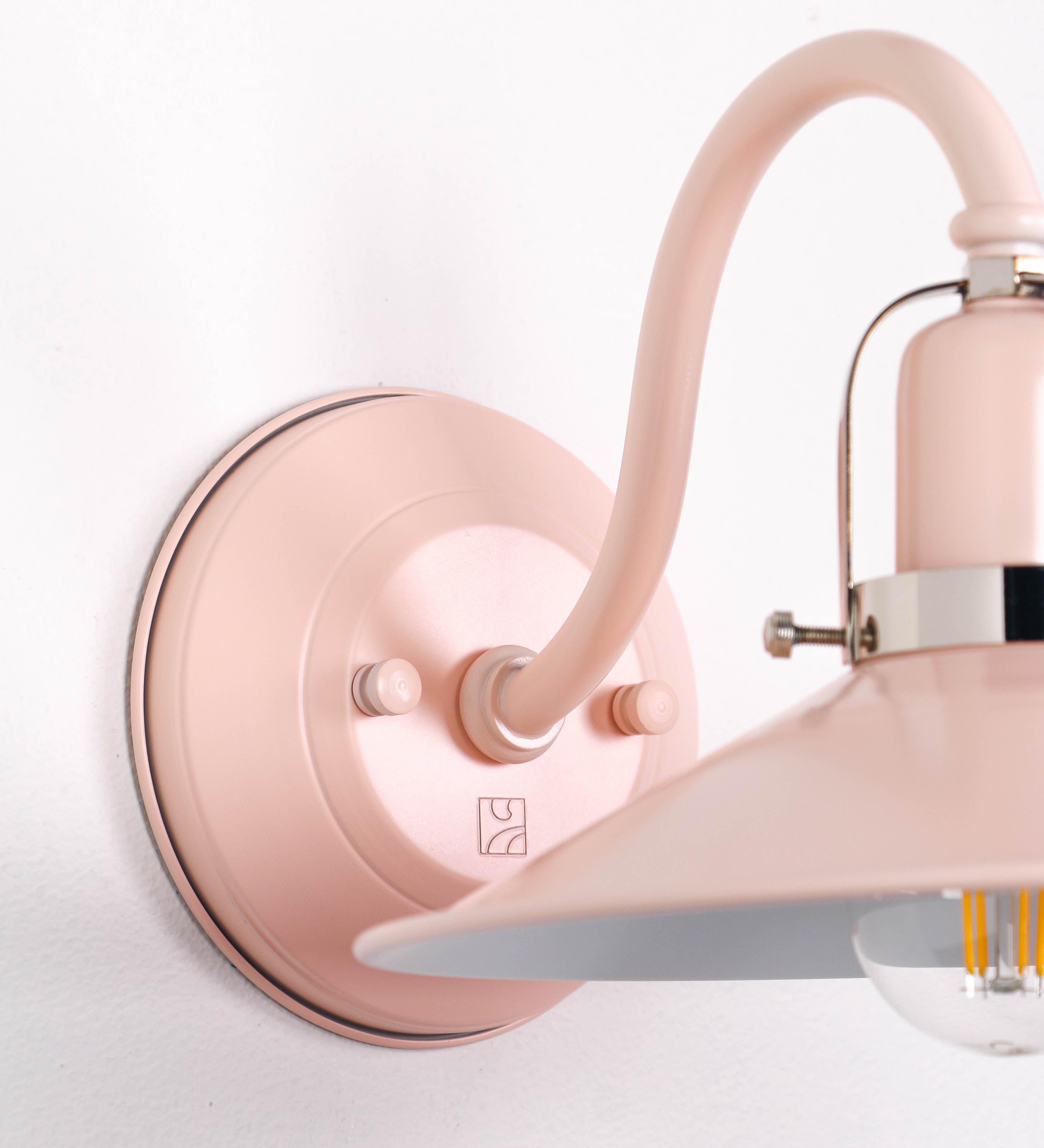 Aplique de parede com um design clássico e cores contemporâneas. O aplique é composto por uma base circular rosa clara, de onde se estende um braço curvo, que forma um arco gracioso e sustenta o chapéu do candeeiro, rosa claro também. O chapéu tem um formato cónico invertido, com uma borda ligeiramente levantada, que direciona a luz para baixo. O interior do chapéu é branco. No topo do chapéu, há um suporte circular em níquel polido, com três braços que fixa o chapéu ao braço curvo.