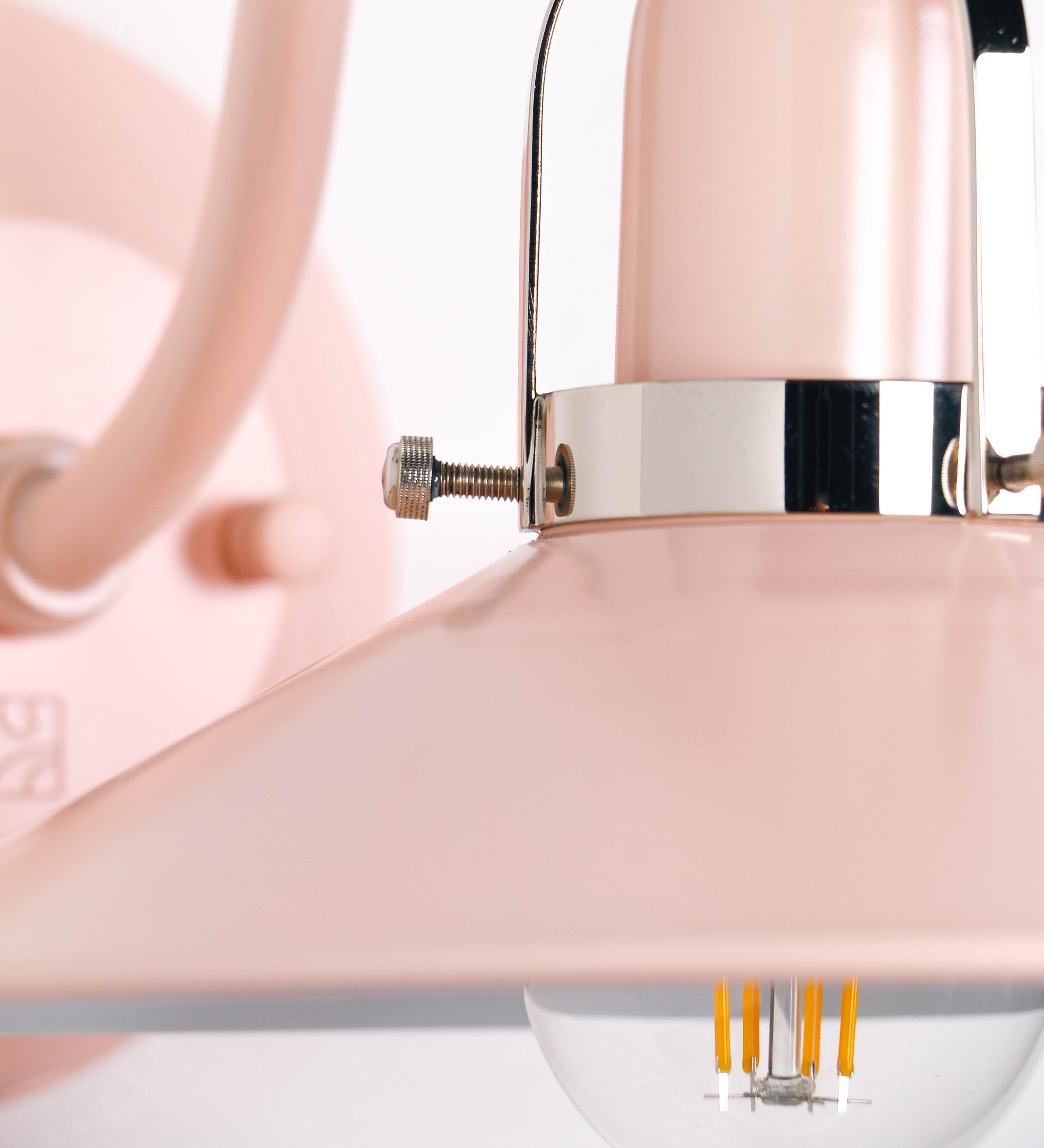 Aplique de parede com um design clássico e cores contemporâneas. O aplique é composto por uma base circular rosa clara, de onde se estende um braço curvo, que forma um arco gracioso e sustenta o chapéu do candeeiro, rosa claro também. O chapéu tem um formato cónico invertido, com uma borda ligeiramente levantada, que direciona a luz para baixo. O interior do chapéu é branco. No topo do chapéu, há um suporte circular em níquel polido, com três braços que fixa o chapéu ao braço curvo.