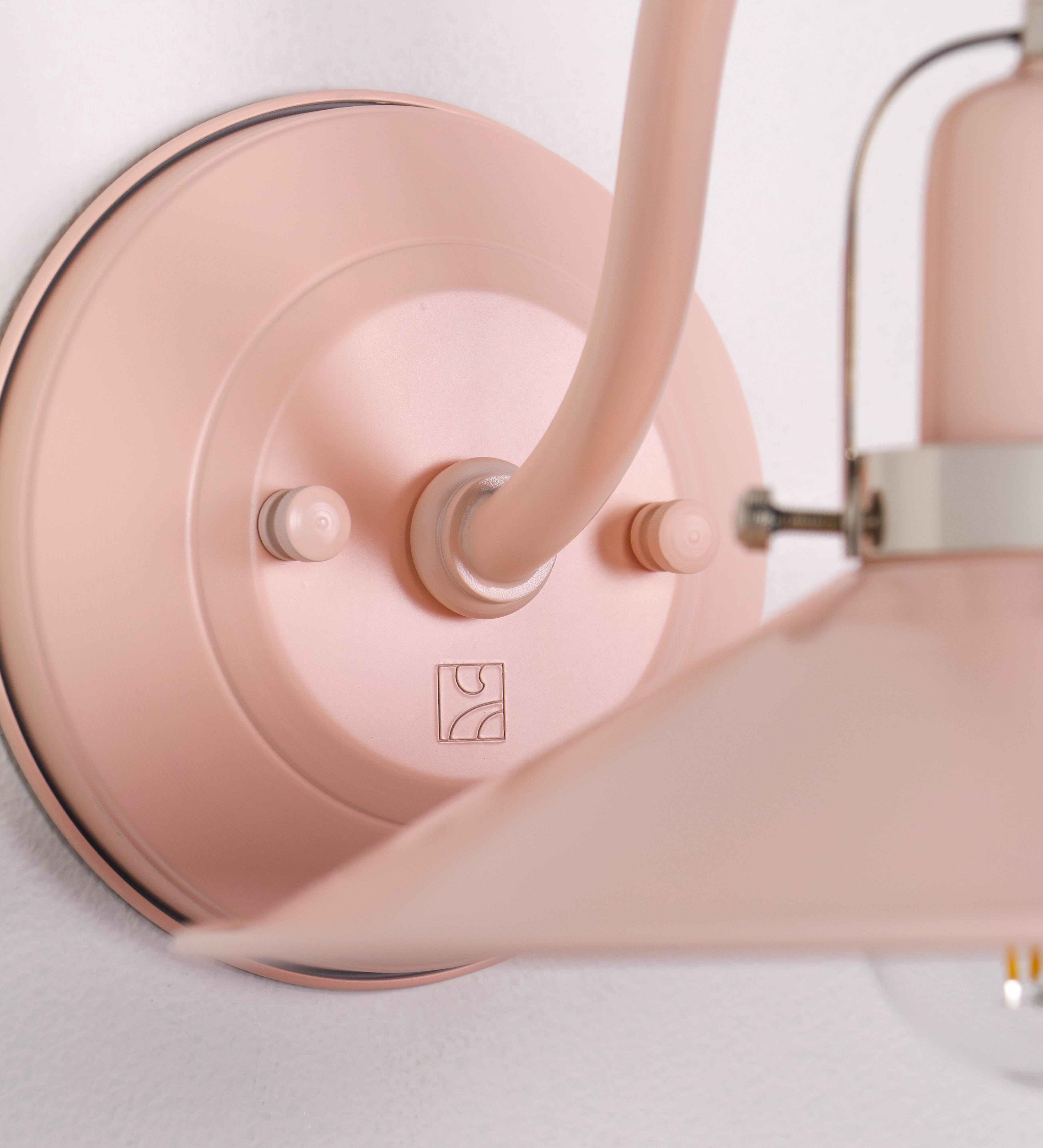 Aplique de parede com um design clássico e cores contemporâneas. O aplique é composto por uma base circular rosa clara, de onde se estende um braço curvo, que forma um arco gracioso e sustenta o chapéu do candeeiro, rosa claro também. O chapéu tem um formato cónico invertido, com uma borda ligeiramente levantada, que direciona a luz para baixo. O interior do chapéu é branco. No topo do chapéu, há um suporte circular em níquel polido, com três braços que fixa o chapéu ao braço curvo.