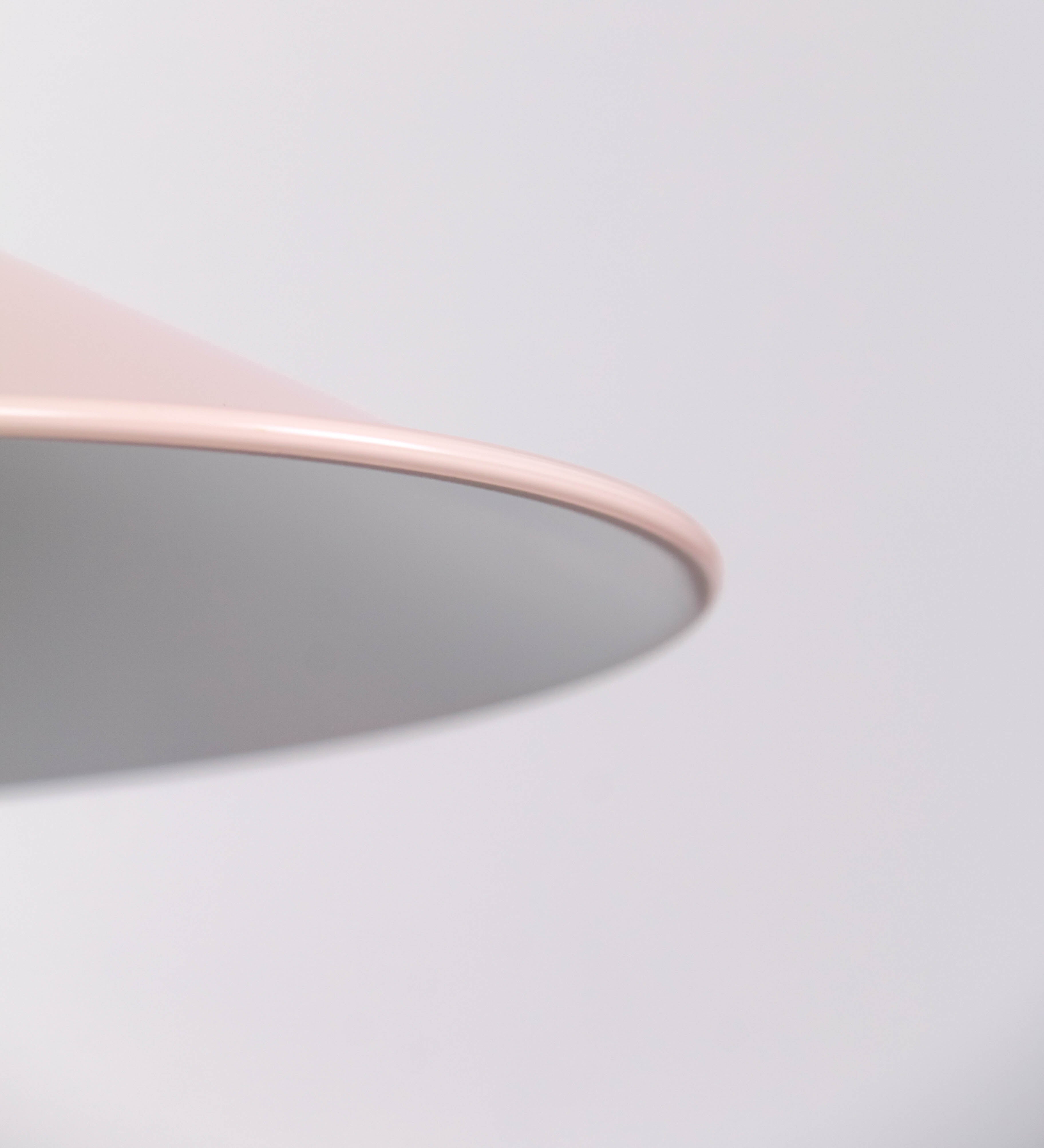 Aplique de parede com um design clássico e cores contemporâneas. O aplique é composto por uma base circular rosa clara, de onde se estende um braço curvo, que forma um arco gracioso e sustenta o chapéu do candeeiro, rosa claro também. O chapéu tem um formato cónico invertido, com uma borda ligeiramente levantada, que direciona a luz para baixo. O interior do chapéu é branco. No topo do chapéu, há um suporte circular em níquel polido, com três braços que fixa o chapéu ao braço curvo.