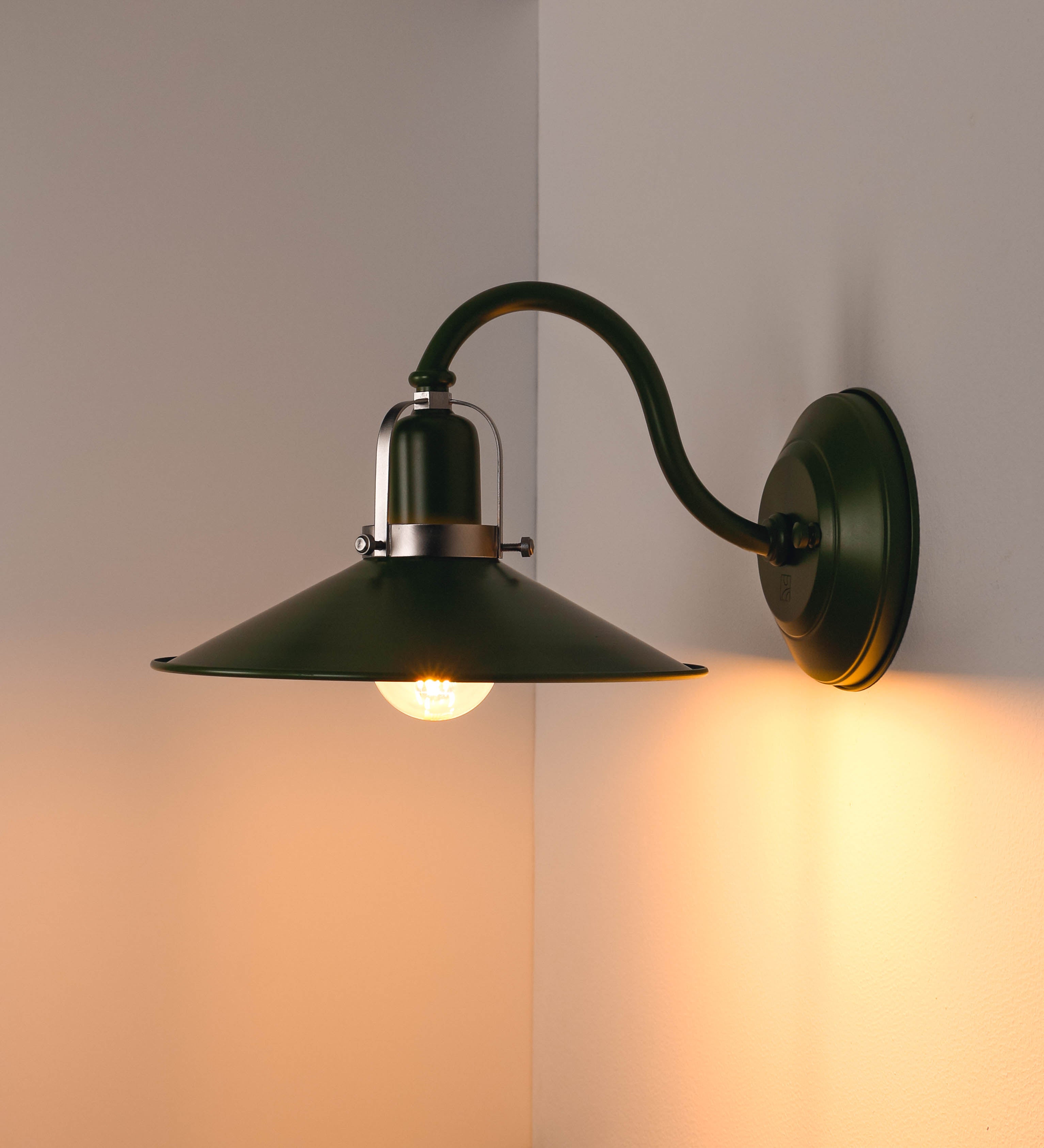 Aplique de parede com um design clássico e cores contemporâneas. O aplique é composto por uma base circular verde couve-de-bruxelas, de onde se estende um braço curvo, que forma um arco gracioso e sustenta o chapéu do candeeiro, verde também. O chapéu tem um formato cónico invertido, com uma borda ligeiramente levantada, que direciona a luz para baixo. O interior do chapéu é branco. No topo do chapéu, há um suporte circular em níquel escovado, com três braços que fixa o chapéu ao braço curvo.
