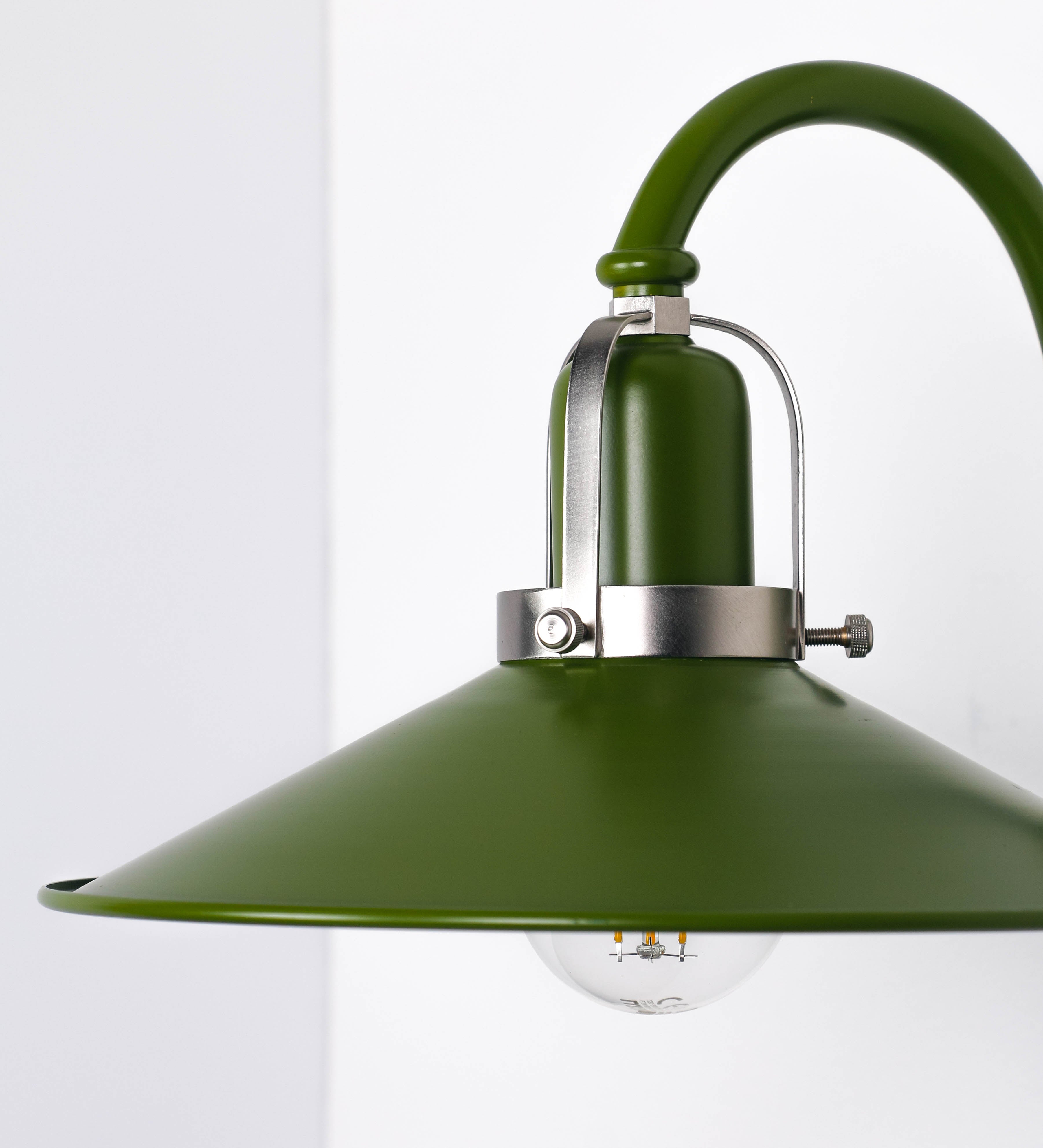 Aplique de parede com um design clássico e cores contemporâneas. O aplique é composto por uma base circular verde couve-de-bruxelas, de onde se estende um braço curvo, que forma um arco gracioso e sustenta o chapéu do candeeiro, verde também. O chapéu tem um formato cónico invertido, com uma borda ligeiramente levantada, que direciona a luz para baixo. O interior do chapéu é branco. No topo do chapéu, há um suporte circular em níquel escovado, com três braços que fixa o chapéu ao braço curvo.