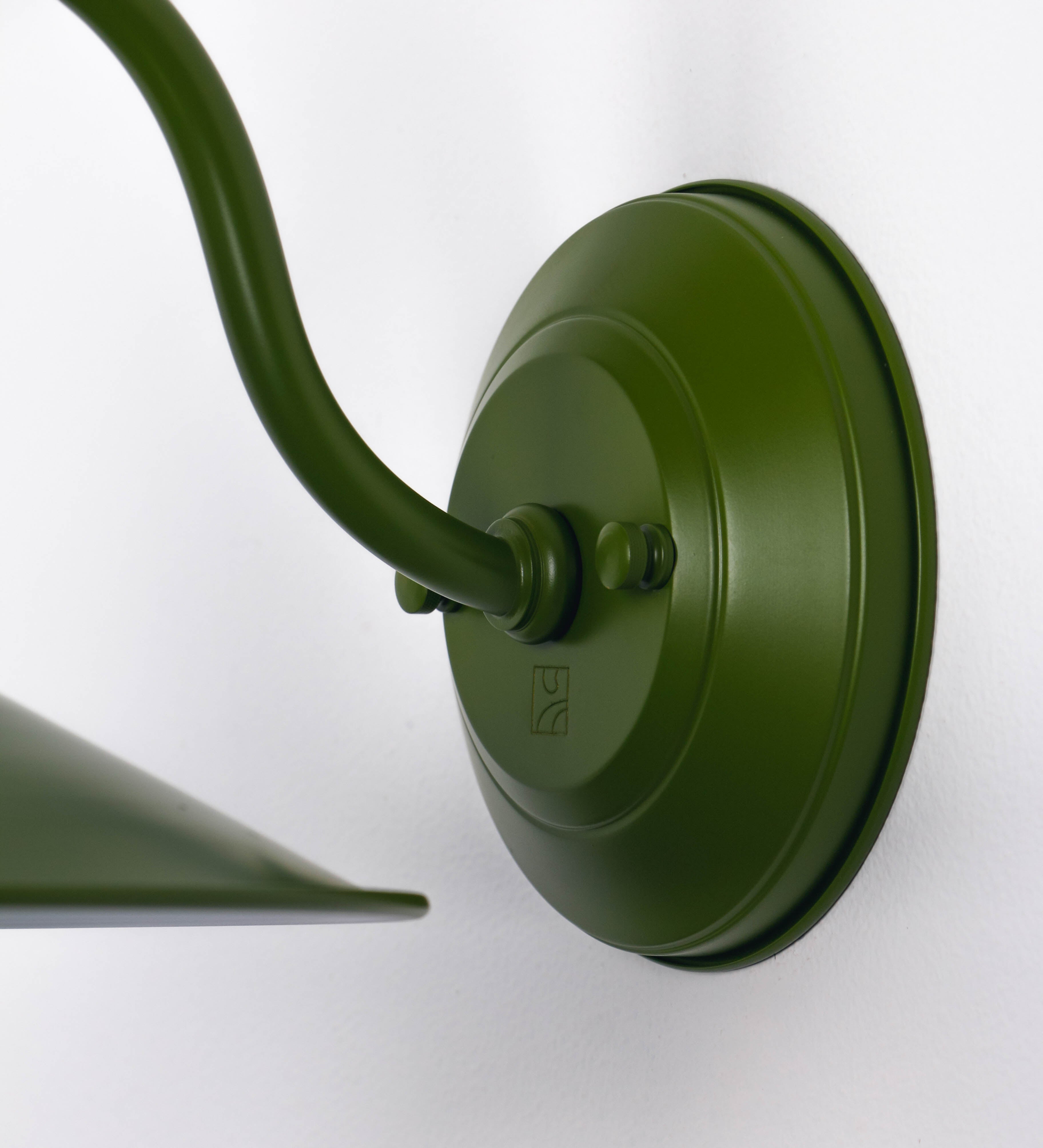 Aplique de parede com um design clássico e cores contemporâneas. O aplique é composto por uma base circular verde couve-de-bruxelas, de onde se estende um braço curvo, que forma um arco gracioso e sustenta o chapéu do candeeiro, verde também. O chapéu tem um formato cónico invertido, com uma borda ligeiramente levantada, que direciona a luz para baixo. O interior do chapéu é branco. No topo do chapéu, há um suporte circular em níquel escovado, com três braços que fixa o chapéu ao braço curvo.