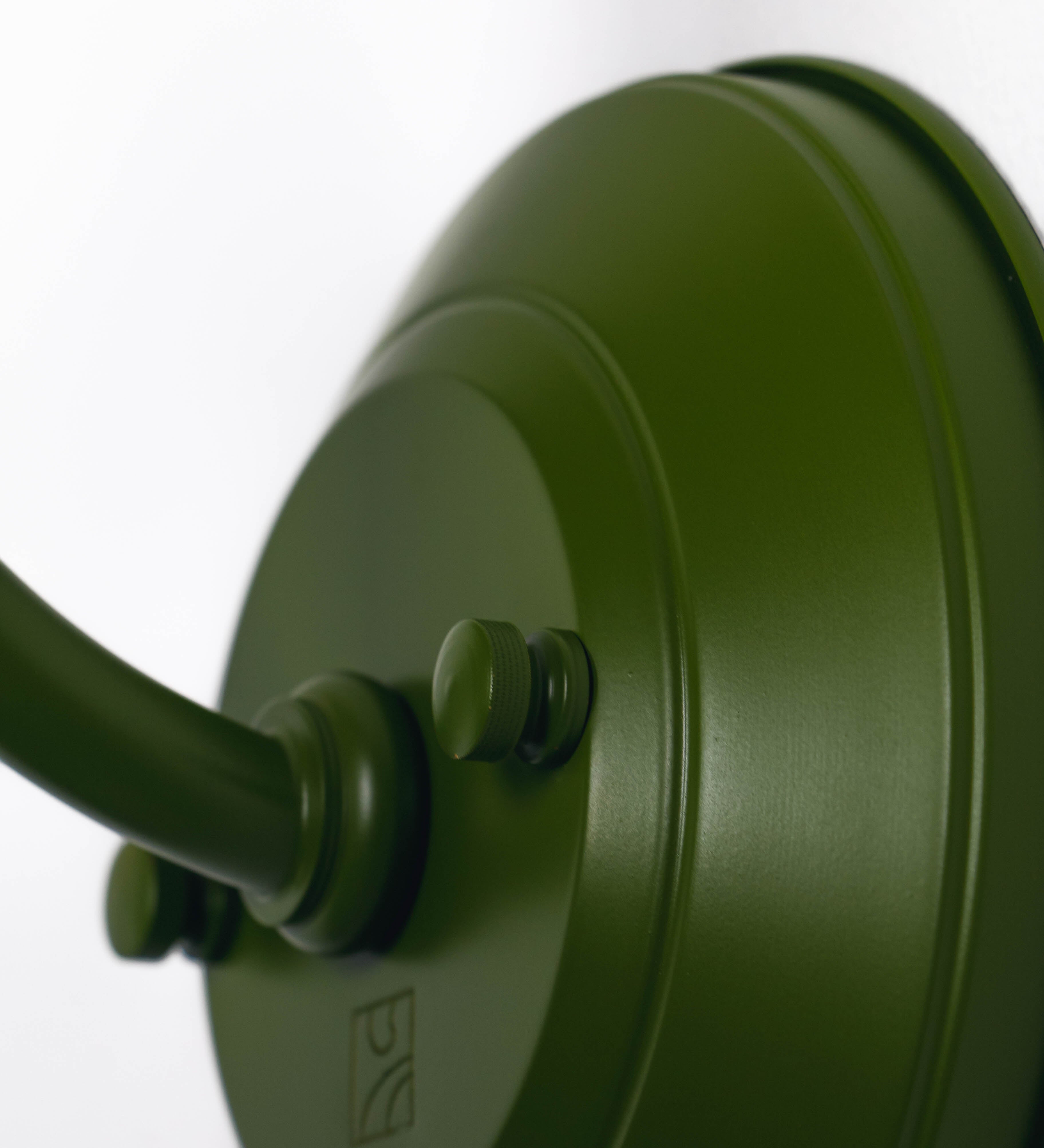 Aplique de parede com um design clássico e cores contemporâneas. O aplique é composto por uma base circular verde couve-de-bruxelas, de onde se estende um braço curvo, que forma um arco gracioso e sustenta o chapéu do candeeiro, verde também. O chapéu tem um formato cónico invertido, com uma borda ligeiramente levantada, que direciona a luz para baixo. O interior do chapéu é branco. No topo do chapéu, há um suporte circular em níquel escovado, com três braços que fixa o chapéu ao braço curvo.