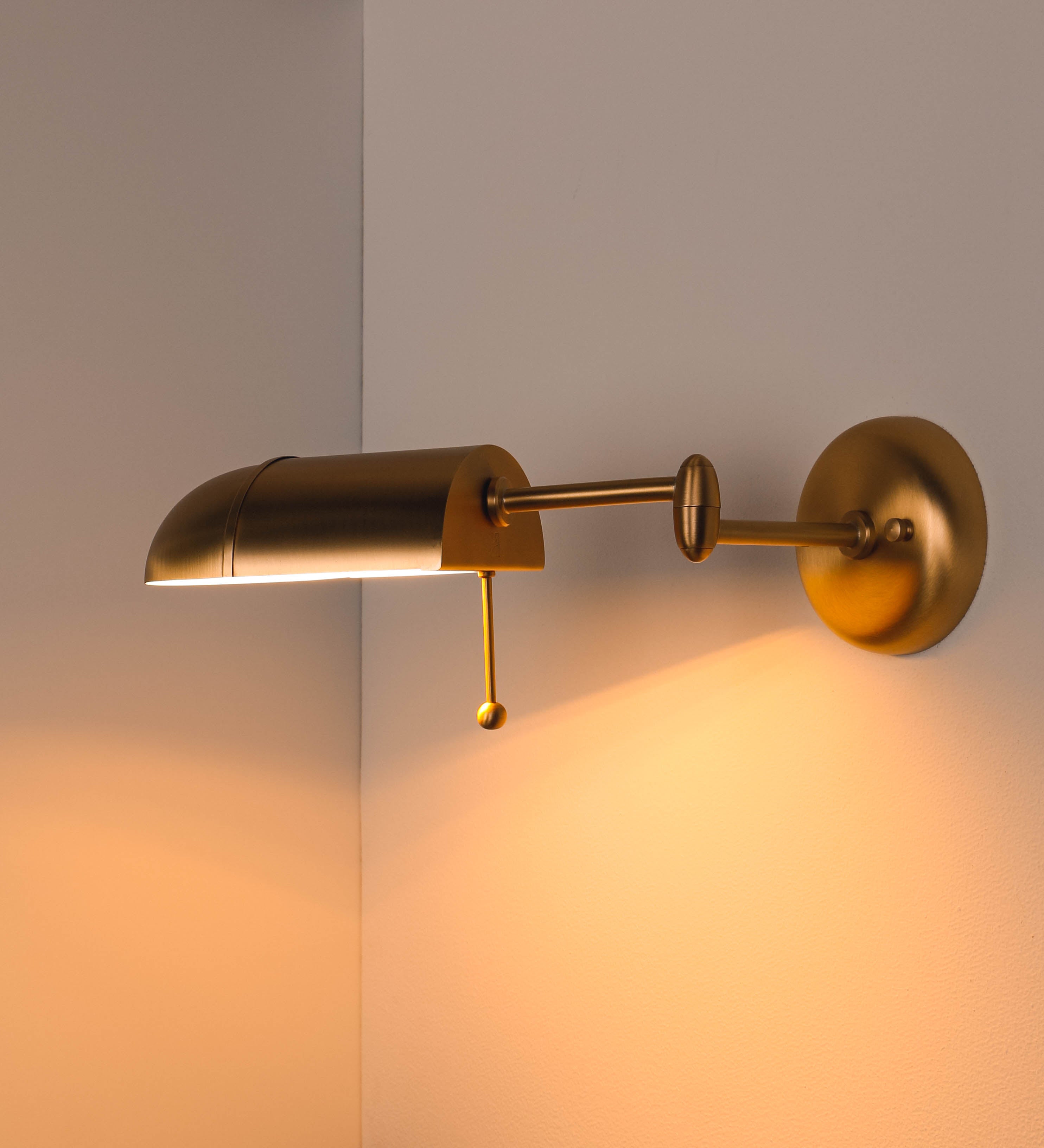 Aplique de parede com um design elegante e funcional. O aplique é todo em latão escovado. A base do aplique é redonda e fixa-se diretamente na parede. Do centro da sua base sai o braço do aplique, que é reto e horizontal. O braço é ajustável permitindo direcionar a luz. No final do braço, há um chapéu longo e arredondado e com uma pequena haste com uma esfera na ponta, que auxilia a movimentar o chapéu. O interior do chapéu é branco.