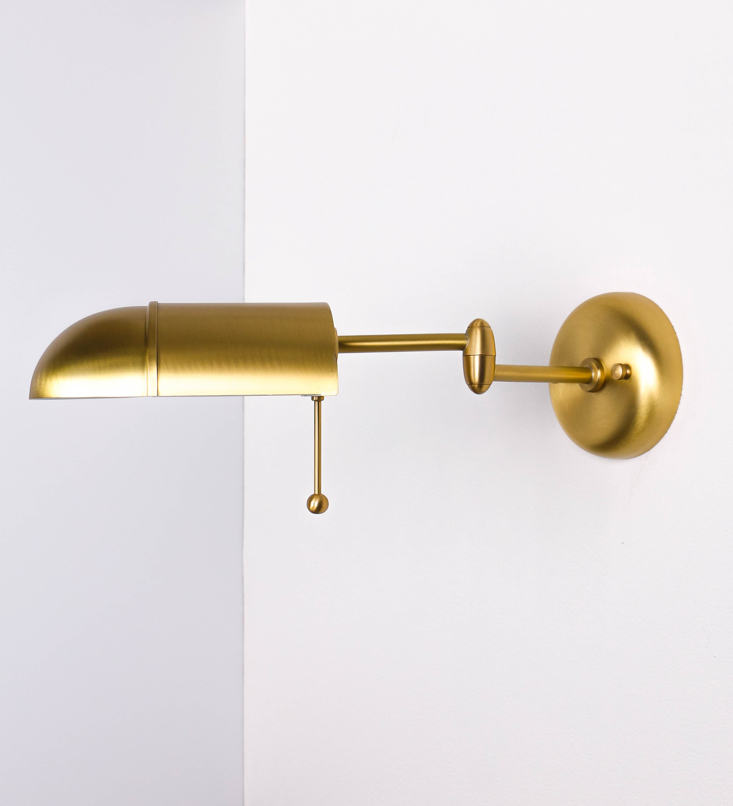 Aplique de parede com um design elegante e funcional. O aplique é todo em latão escovado. A base do aplique é redonda e fixa-se diretamente na parede. Do centro da sua base sai o braço do aplique, que é reto e horizontal. O braço é ajustável permitindo direcionar a luz. No final do braço, há um chapéu longo e arredondado e com uma pequena haste com uma esfera na ponta, que auxilia a movimentar o chapéu. O interior do chapéu é branco.