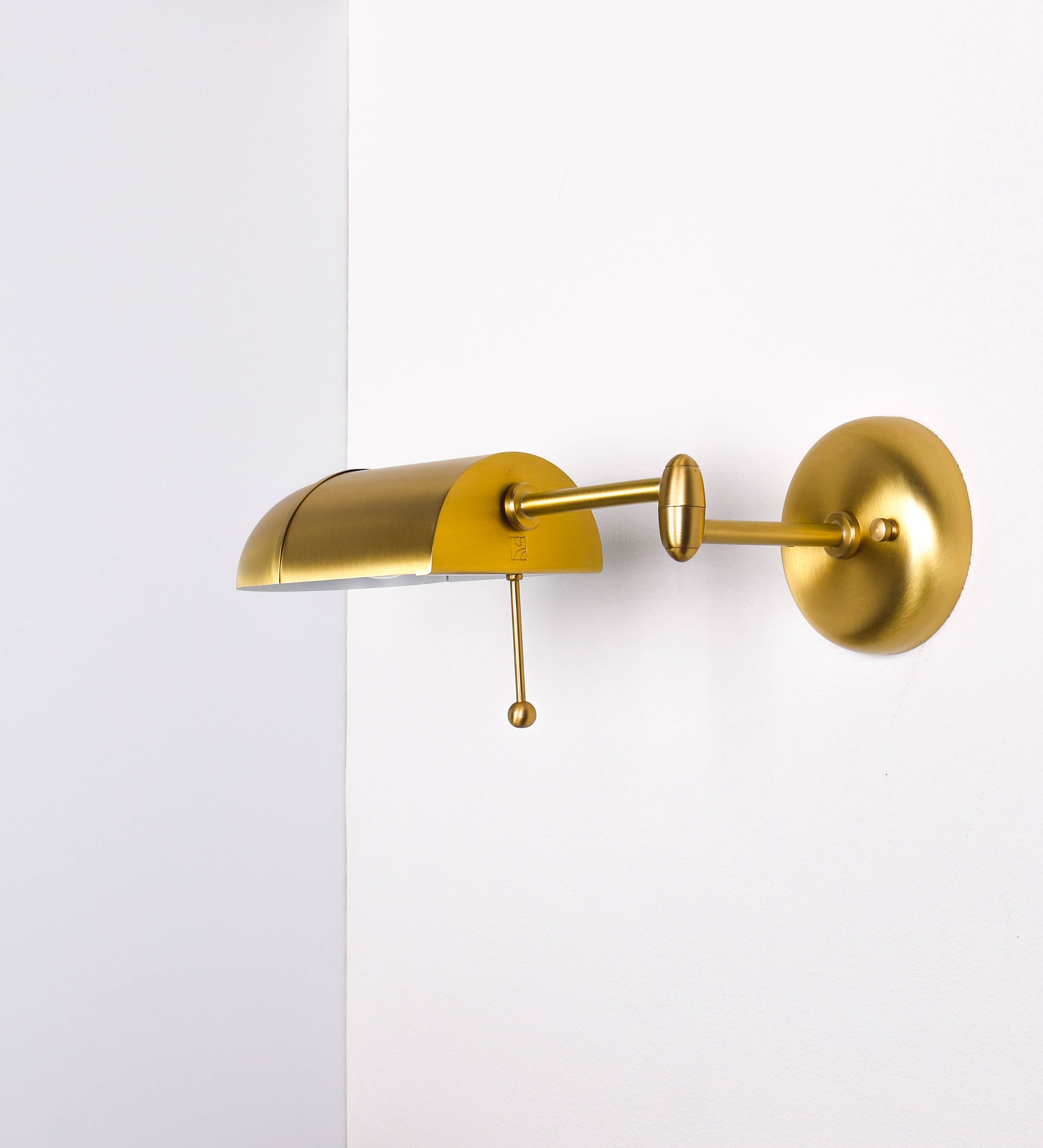 Aplique de parede com um design elegante e funcional. O aplique é todo em latão escovado. A base do aplique é redonda e fixa-se diretamente na parede. Do centro da sua base sai o braço do aplique, que é reto e horizontal. O braço é ajustável permitindo direcionar a luz. No final do braço, há um chapéu longo e arredondado e com uma pequena haste com uma esfera na ponta, que auxilia a movimentar o chapéu. O interior do chapéu é branco.