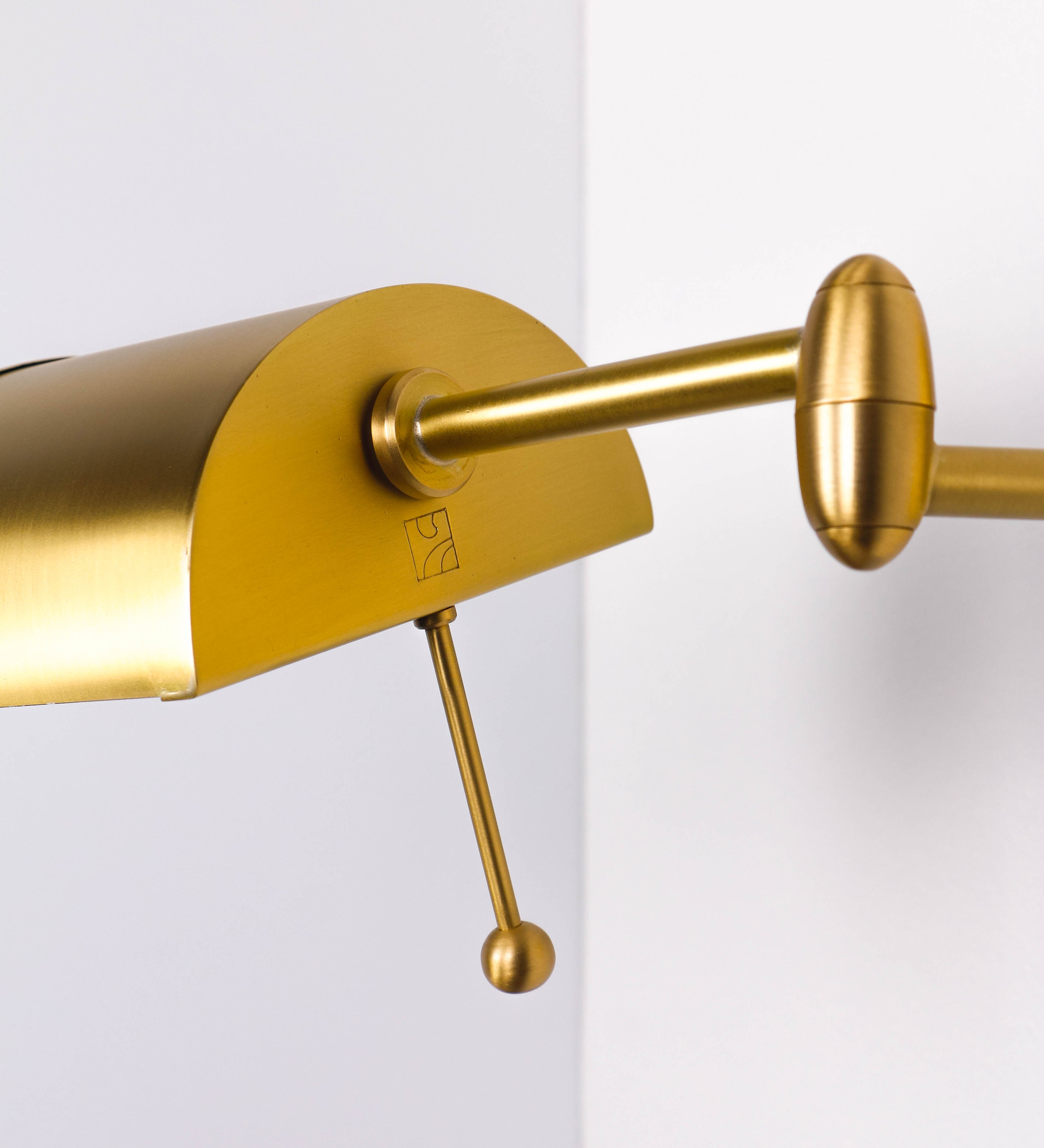 Aplique de parede com um design elegante e funcional. O aplique é todo em latão escovado. A base do aplique é redonda e fixa-se diretamente na parede. Do centro da sua base sai o braço do aplique, que é reto e horizontal. O braço é ajustável permitindo direcionar a luz. No final do braço, há um chapéu longo e arredondado e com uma pequena haste com uma esfera na ponta, que auxilia a movimentar o chapéu. O interior do chapéu é branco.