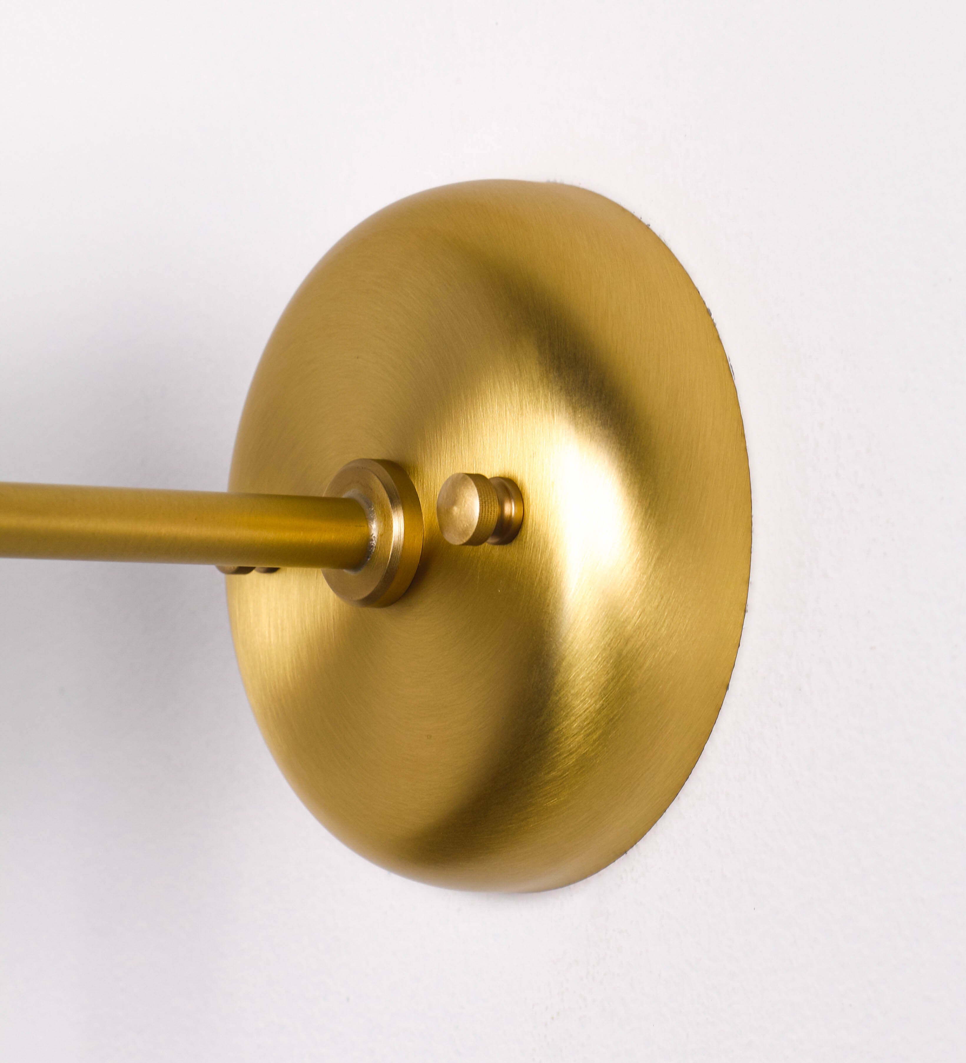Aplique de parede com um design elegante e funcional. O aplique é todo em latão escovado. A base do aplique é redonda e fixa-se diretamente na parede. Do centro da sua base sai o braço do aplique, que é reto e horizontal. O braço é ajustável permitindo direcionar a luz. No final do braço, há um chapéu longo e arredondado e com uma pequena haste com uma esfera na ponta, que auxilia a movimentar o chapéu. O interior do chapéu é branco.