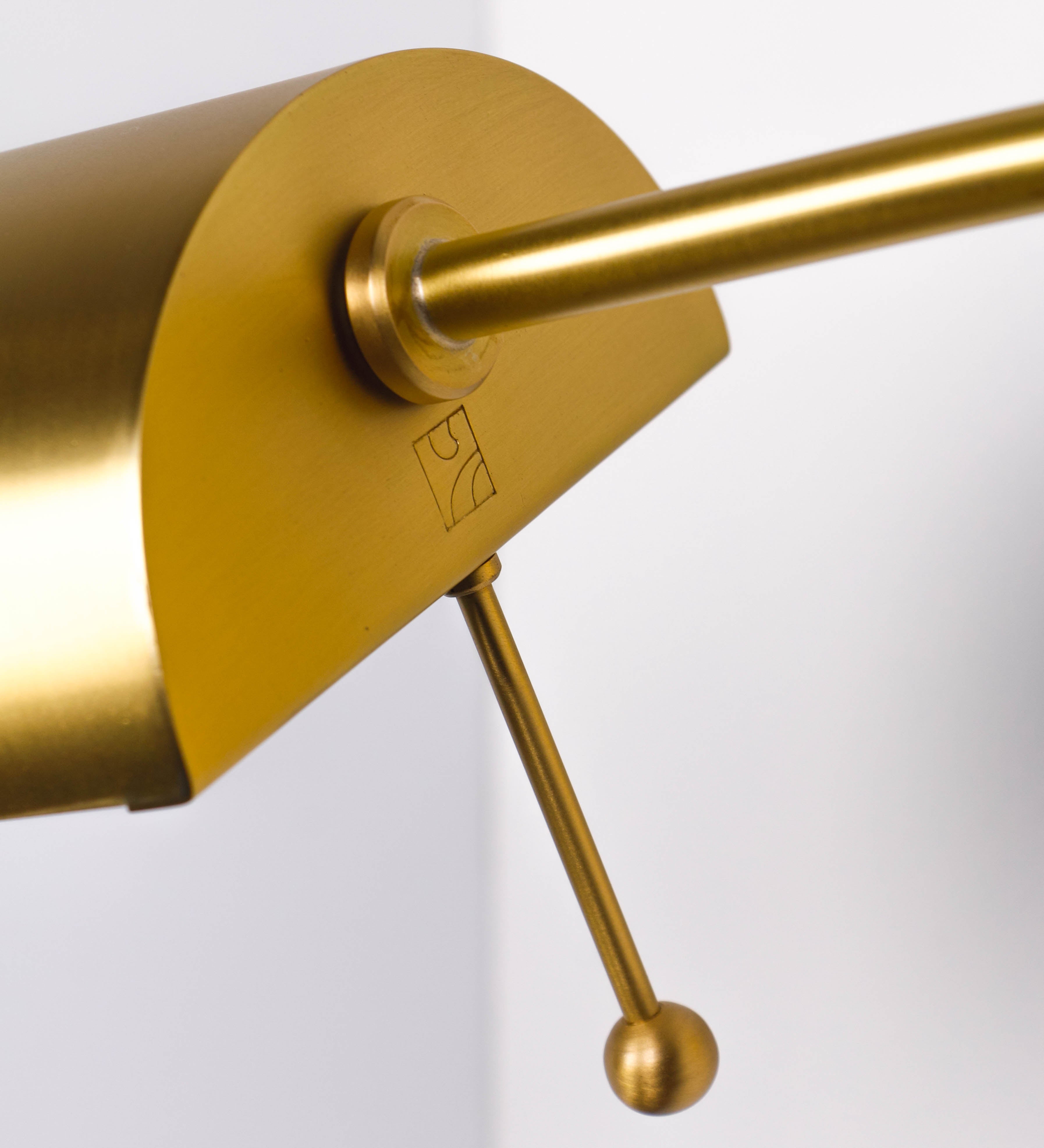 Aplique de parede com um design elegante e funcional. O aplique é todo em latão escovado. A base do aplique é redonda e fixa-se diretamente na parede. Do centro da sua base sai o braço do aplique, que é reto e horizontal. O braço é ajustável permitindo direcionar a luz. No final do braço, há um chapéu longo e arredondado e com uma pequena haste com uma esfera na ponta, que auxilia a movimentar o chapéu. O interior do chapéu é branco.