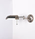 Aplique de parede com um design elegante e funcional. O aplique é todo em níquel escovado. A base do aplique é redonda e fixa-se diretamente na parede. Do centro da sua base sai o braço do aplique, que é reto e horizontal. O braço é ajustável permitindo direcionar a luz. No final do braço, há um chapéu longo e arredondado e com uma pequena haste com uma esfera na ponta, que auxilia a movimentar o chapéu. O interior do chapéu é branco.