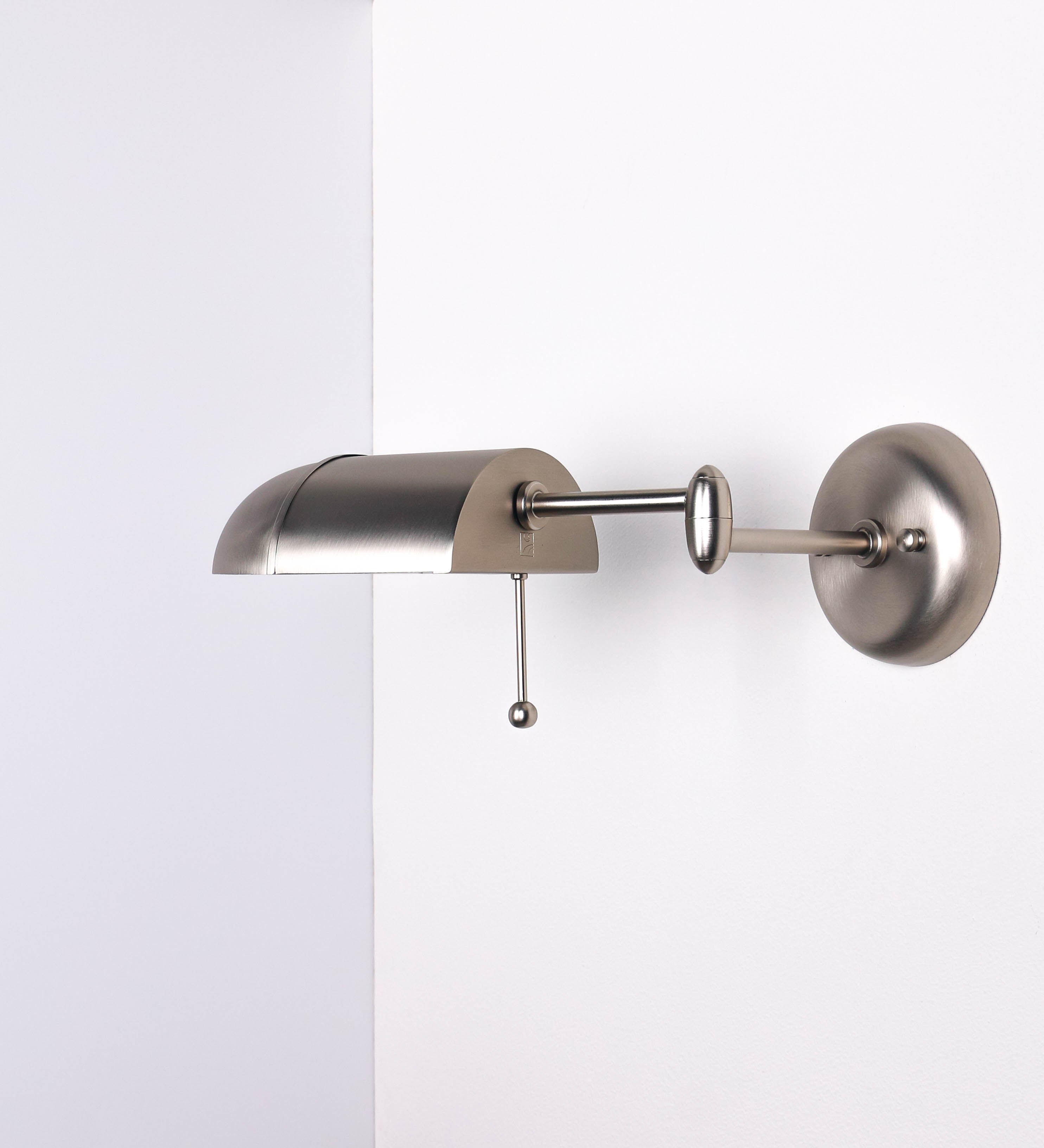 Aplique de parede com um design elegante e funcional. O aplique é todo em níquel escovado. A base do aplique é redonda e fixa-se diretamente na parede. Do centro da sua base sai o braço do aplique, que é reto e horizontal. O braço é ajustável permitindo direcionar a luz. No final do braço, há um chapéu longo e arredondado e com uma pequena haste com uma esfera na ponta, que auxilia a movimentar o chapéu. O interior do chapéu é branco.