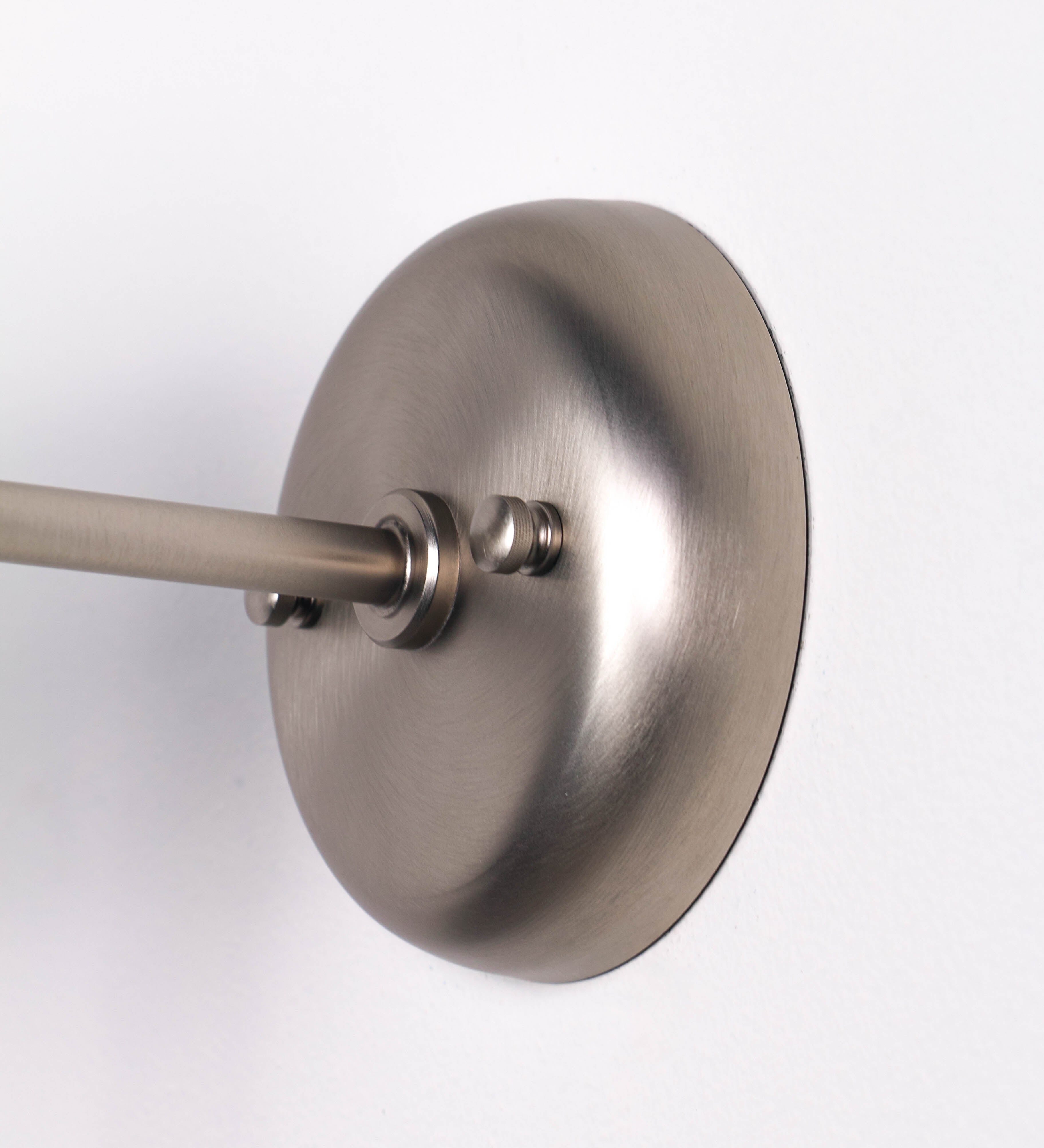 Aplique de parede com um design elegante e funcional. O aplique é todo em níquel escovado. A base do aplique é redonda e fixa-se diretamente na parede. Do centro da sua base sai o braço do aplique, que é reto e horizontal. O braço é ajustável permitindo direcionar a luz. No final do braço, há um chapéu longo e arredondado e com uma pequena haste com uma esfera na ponta, que auxilia a movimentar o chapéu. O interior do chapéu é branco.