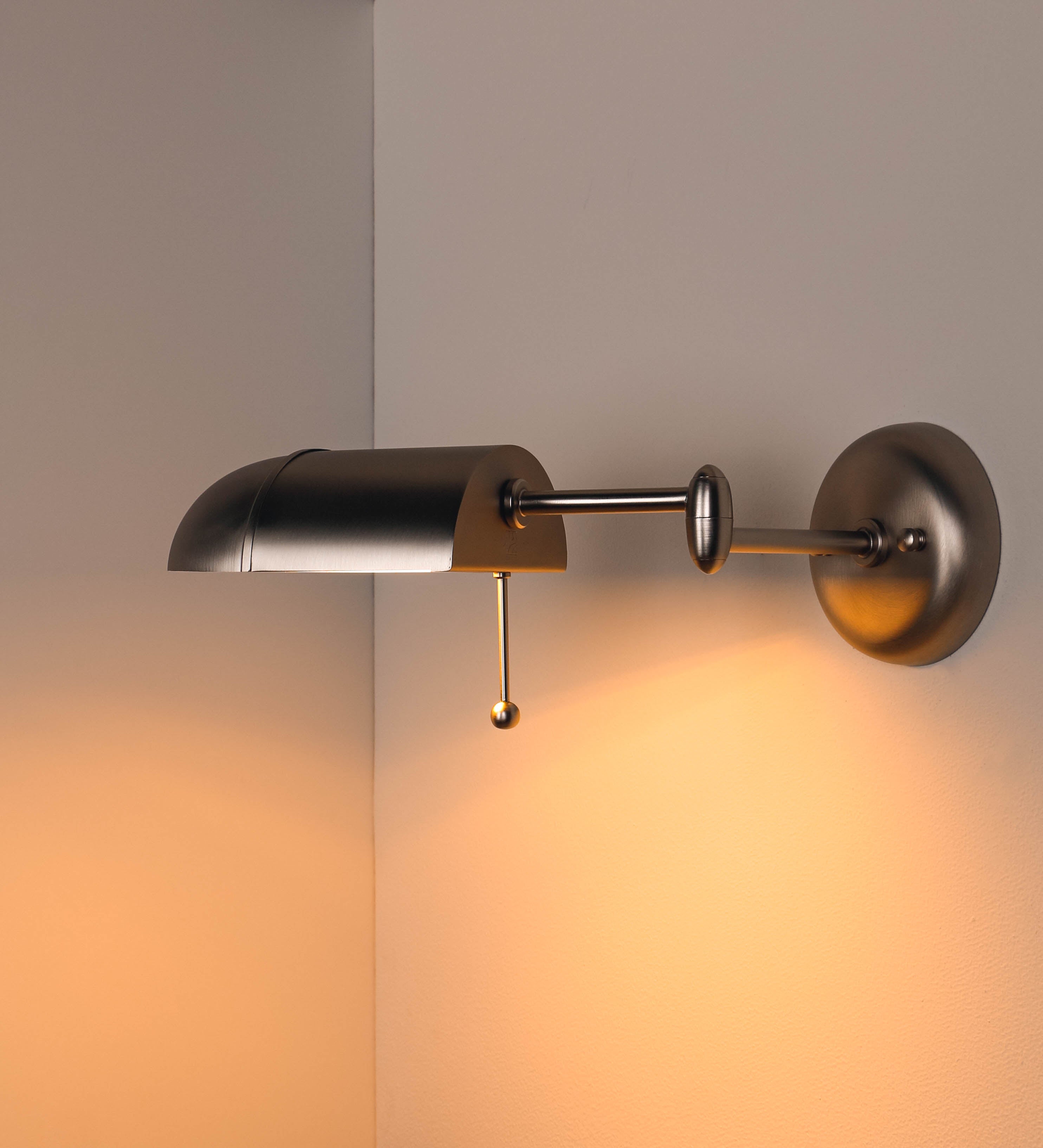 Aplique de parede com um design elegante e funcional. O aplique é todo em níquel escovado. A base do aplique é redonda e fixa-se diretamente na parede. Do centro da sua base sai o braço do aplique, que é reto e horizontal. O braço é ajustável permitindo direcionar a luz. No final do braço, há um chapéu longo e arredondado e com uma pequena haste com uma esfera na ponta, que auxilia a movimentar o chapéu. O interior do chapéu é branco.