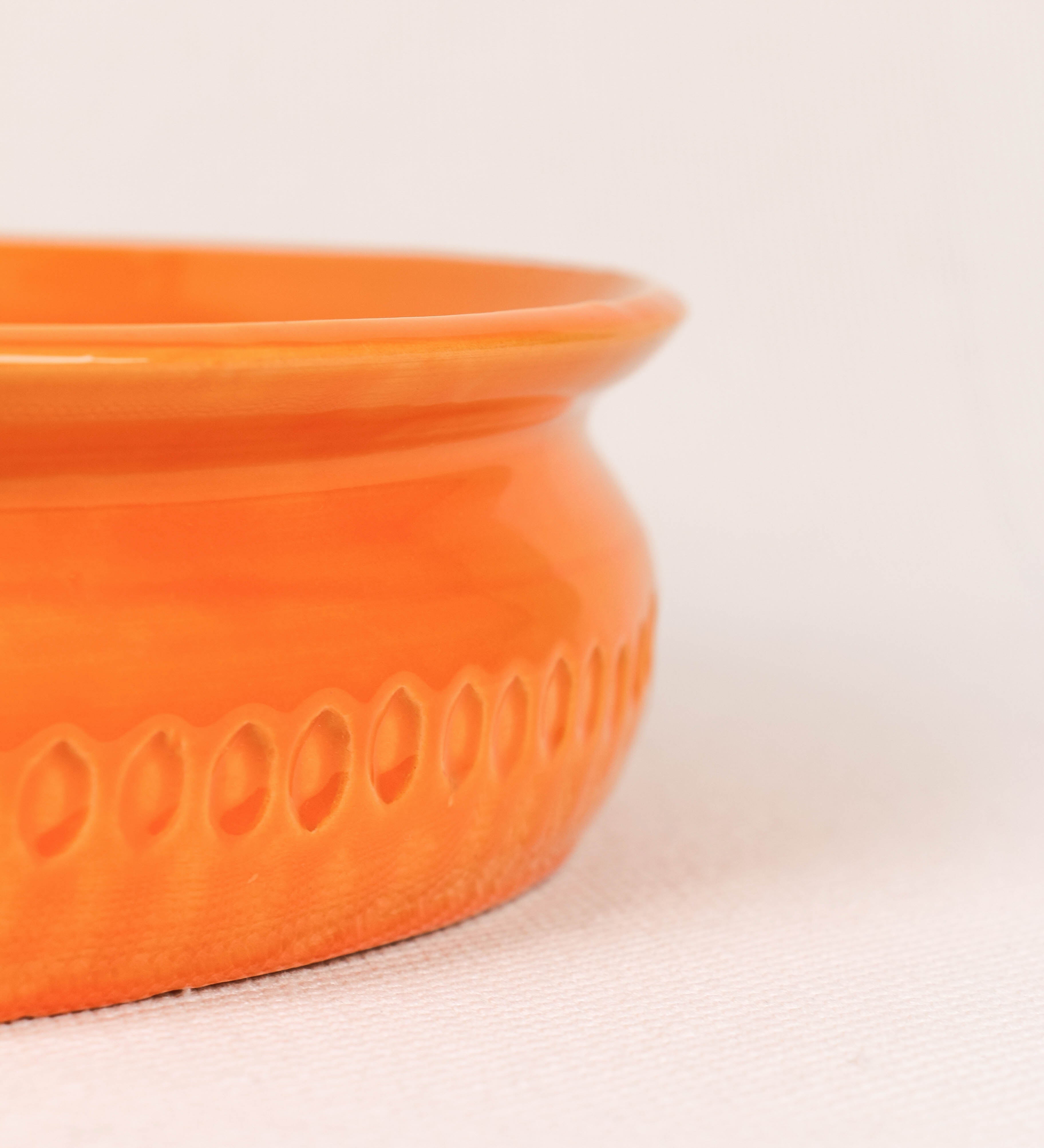 Assadeira com um design rústico e distinto. A assadeira é de barro, vidrado em tom laranja tangerina com brilho, conferindo um caráter chamativo à peça. A forma da assadeira é circular, com uma borda superior mais estreita. A superfície externa da peça é decorada com um padrão de relevos losangulares, que envolvem a assadeira e acrescentam textura e interesse visual ao design.