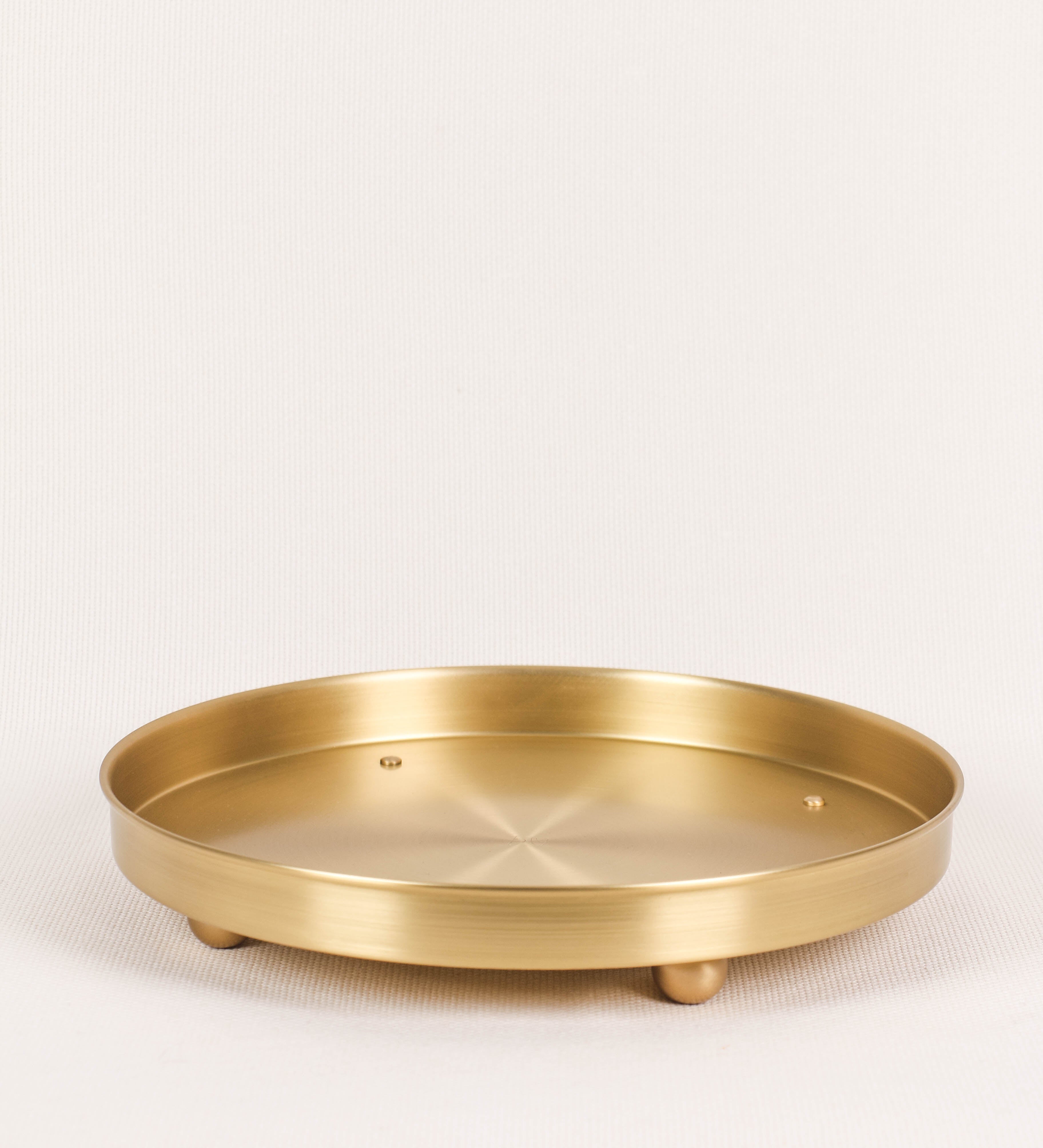 Bandeja com um design sofisticado e intemporal. A bandeja é de latão, com um tom dourado mate e acabamento escovado. Tem uma forma circular, de superfície plana e com uma borda envolvente de altura média e grossura fina. Na sua base tem três pequenas esferas, em latão também, dispostas em formato triangular, que sustentam o prato circular no topo.