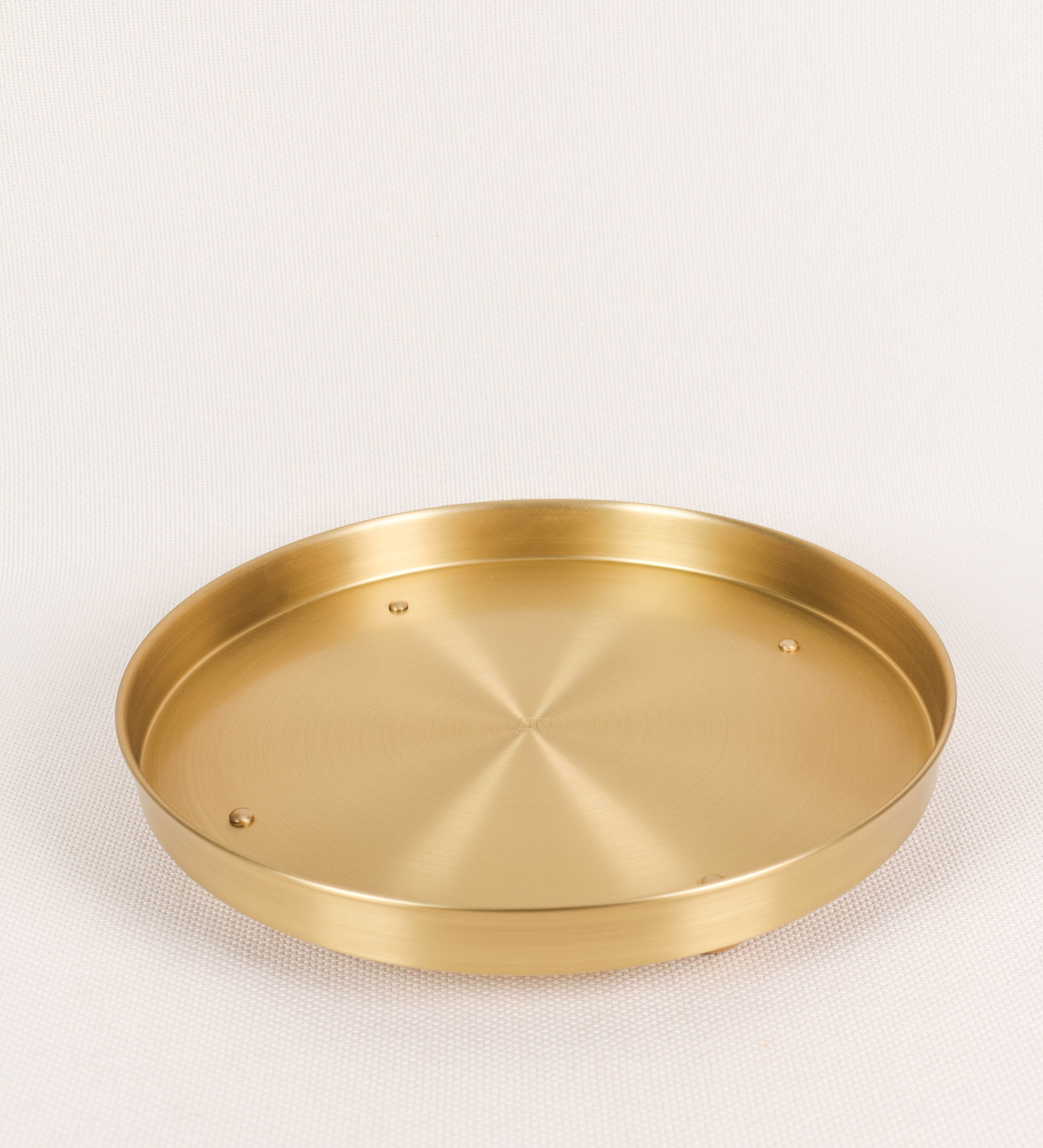 Bandeja com um design sofisticado e intemporal. A bandeja é de latão, com um tom dourado mate e acabamento escovado. Tem uma forma circular, de superfície plana e com uma borda envolvente de altura média e grossura fina. Na sua base tem três pequenas esferas, em latão também, dispostas em formato triangular, que sustentam o prato circular no topo.