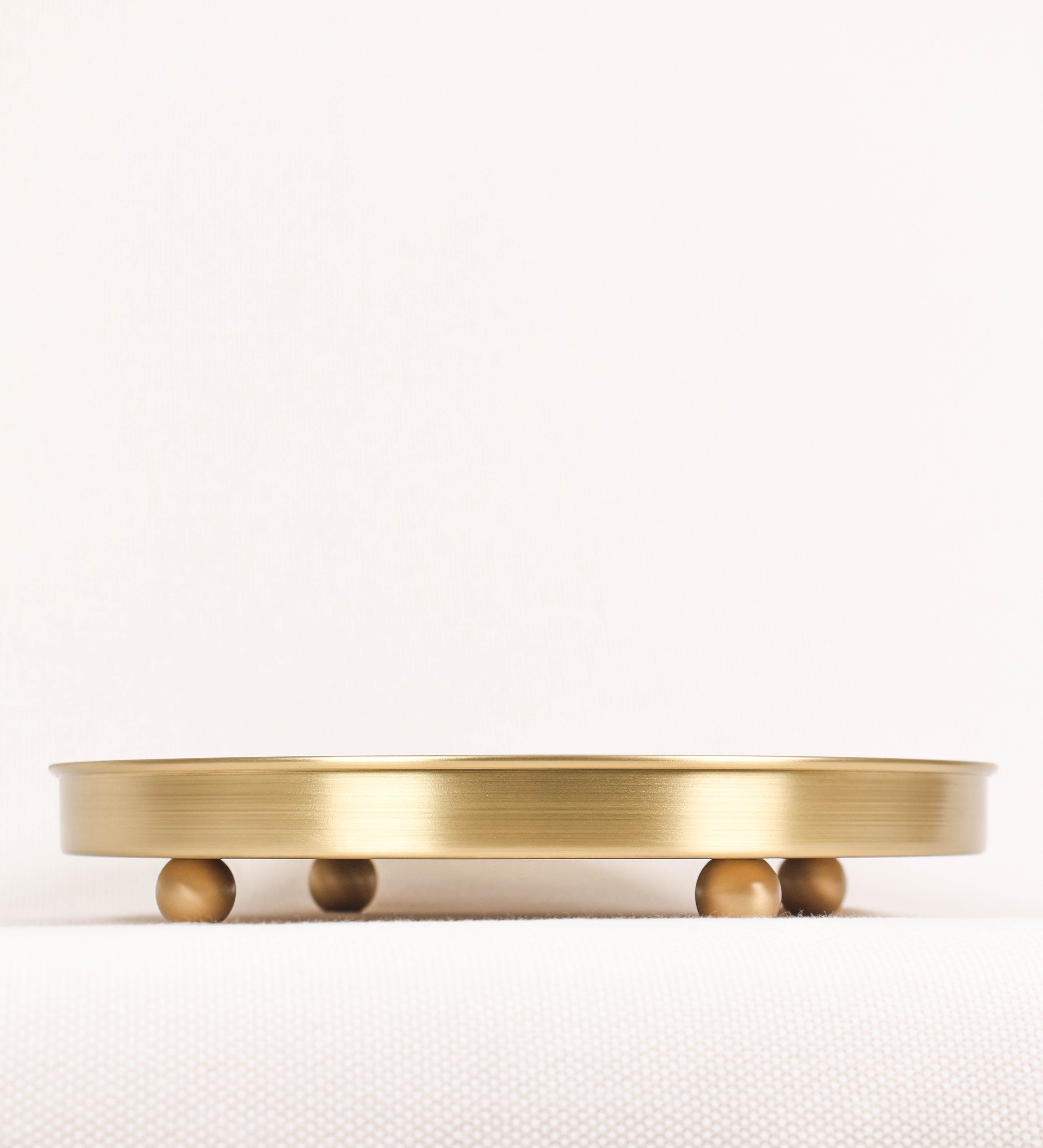 Bandeja com um design sofisticado e intemporal. A bandeja é de latão, com um tom dourado mate e acabamento escovado. Tem uma forma circular, de superfície plana e com uma borda envolvente de altura média e grossura fina. Na sua base tem três pequenas esferas, em latão também, dispostas em formato triangular, que sustentam o prato circular no topo.