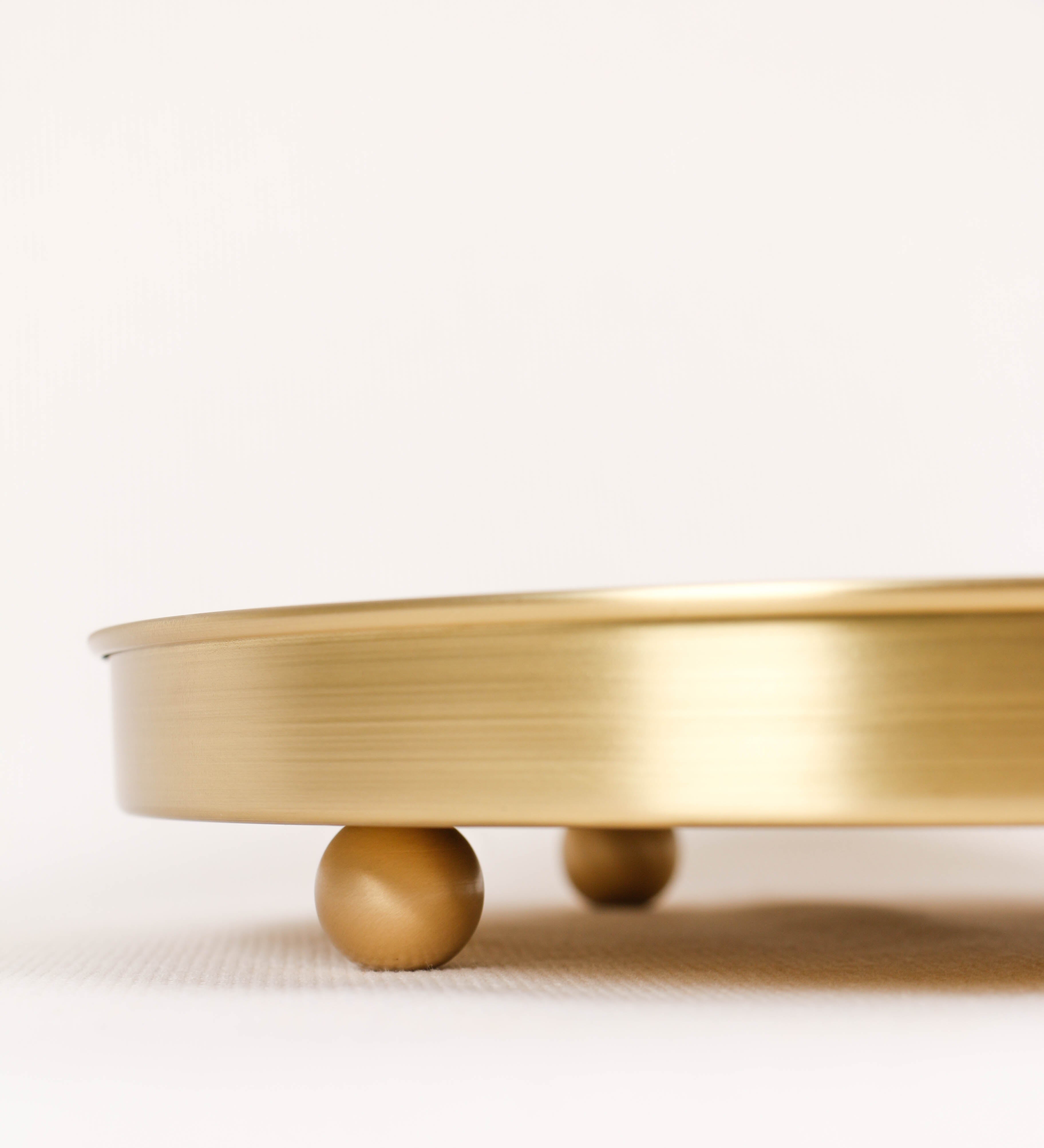 Bandeja com um design sofisticado e intemporal. A bandeja é de latão, com um tom dourado mate e acabamento escovado. Tem uma forma circular, de superfície plana e com uma borda envolvente de altura média e grossura fina. Na sua base tem três pequenas esferas, em latão também, dispostas em formato triangular, que sustentam o prato circular no topo.