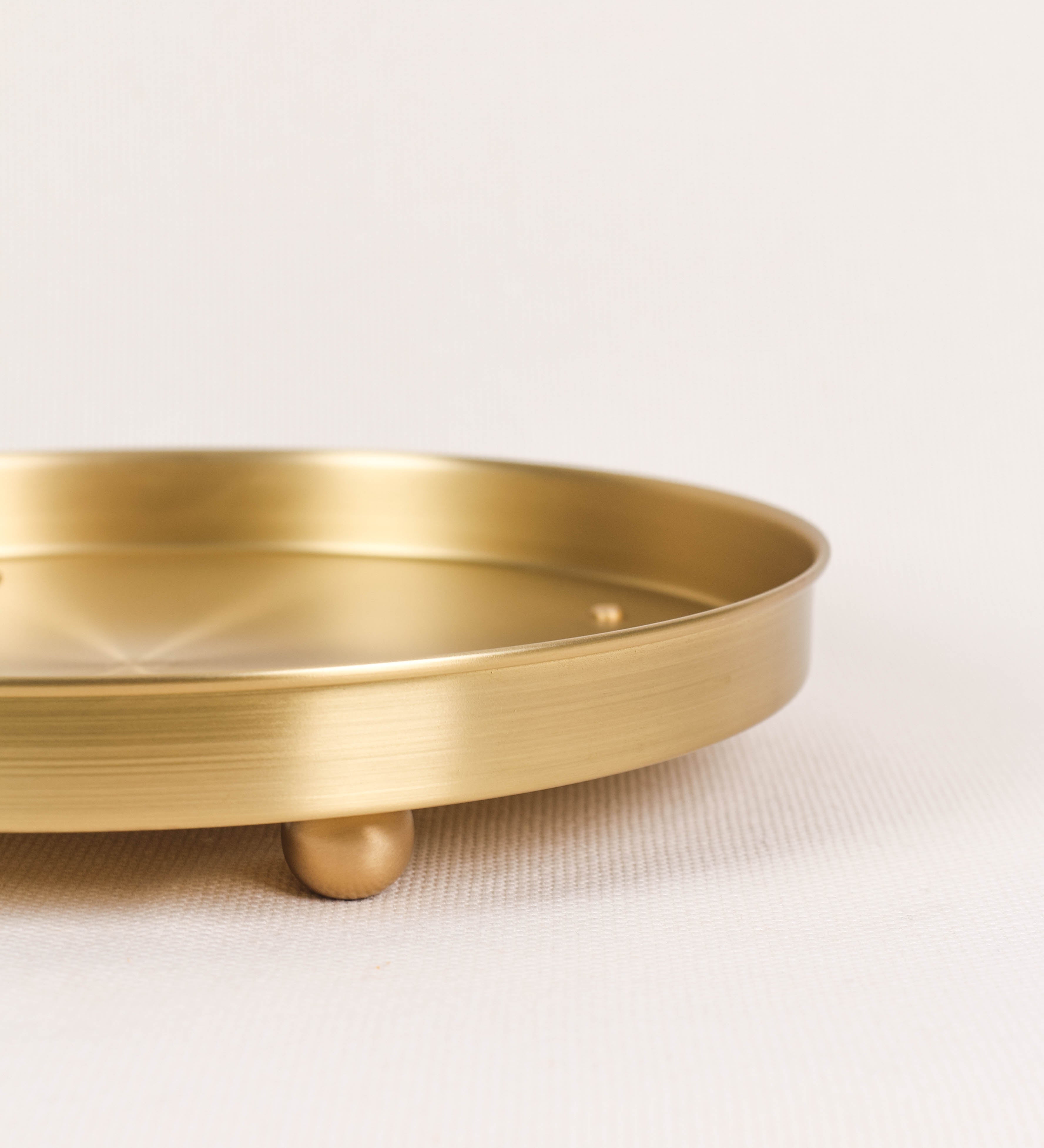 Bandeja com um design sofisticado e intemporal. A bandeja é de latão, com um tom dourado mate e acabamento escovado. Tem uma forma circular, de superfície plana e com uma borda envolvente de altura média e grossura fina. Na sua base tem três pequenas esferas, em latão também, dispostas em formato triangular, que sustentam o prato circular no topo.