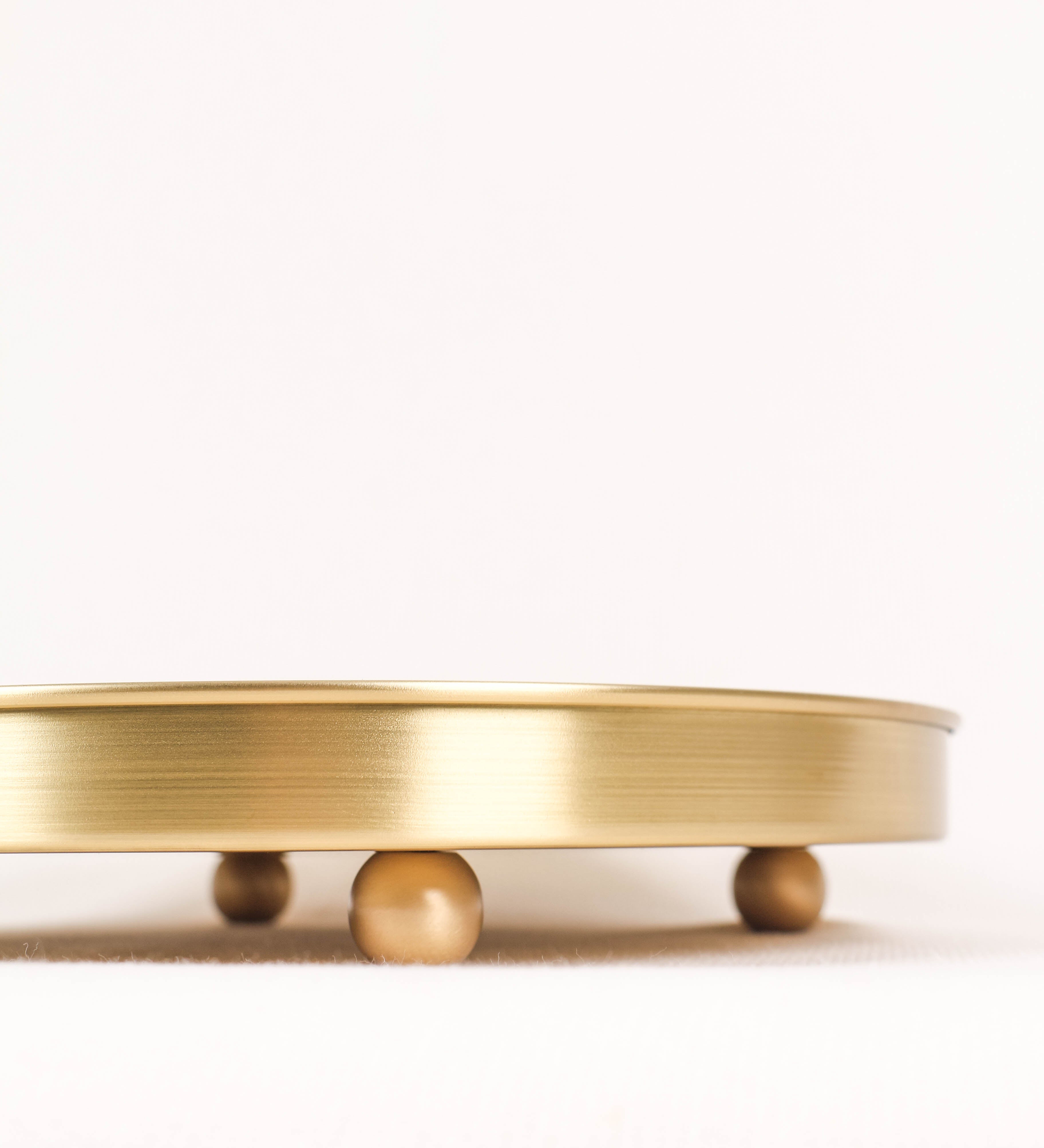 Bandeja com um design sofisticado e intemporal. A bandeja é de latão, com um tom dourado mate e acabamento escovado. Tem uma forma circular, de superfície plana e com uma borda envolvente de altura média e grossura fina. Na sua base tem três pequenas esferas, em latão também, dispostas em formato triangular, que sustentam o prato circular no topo.