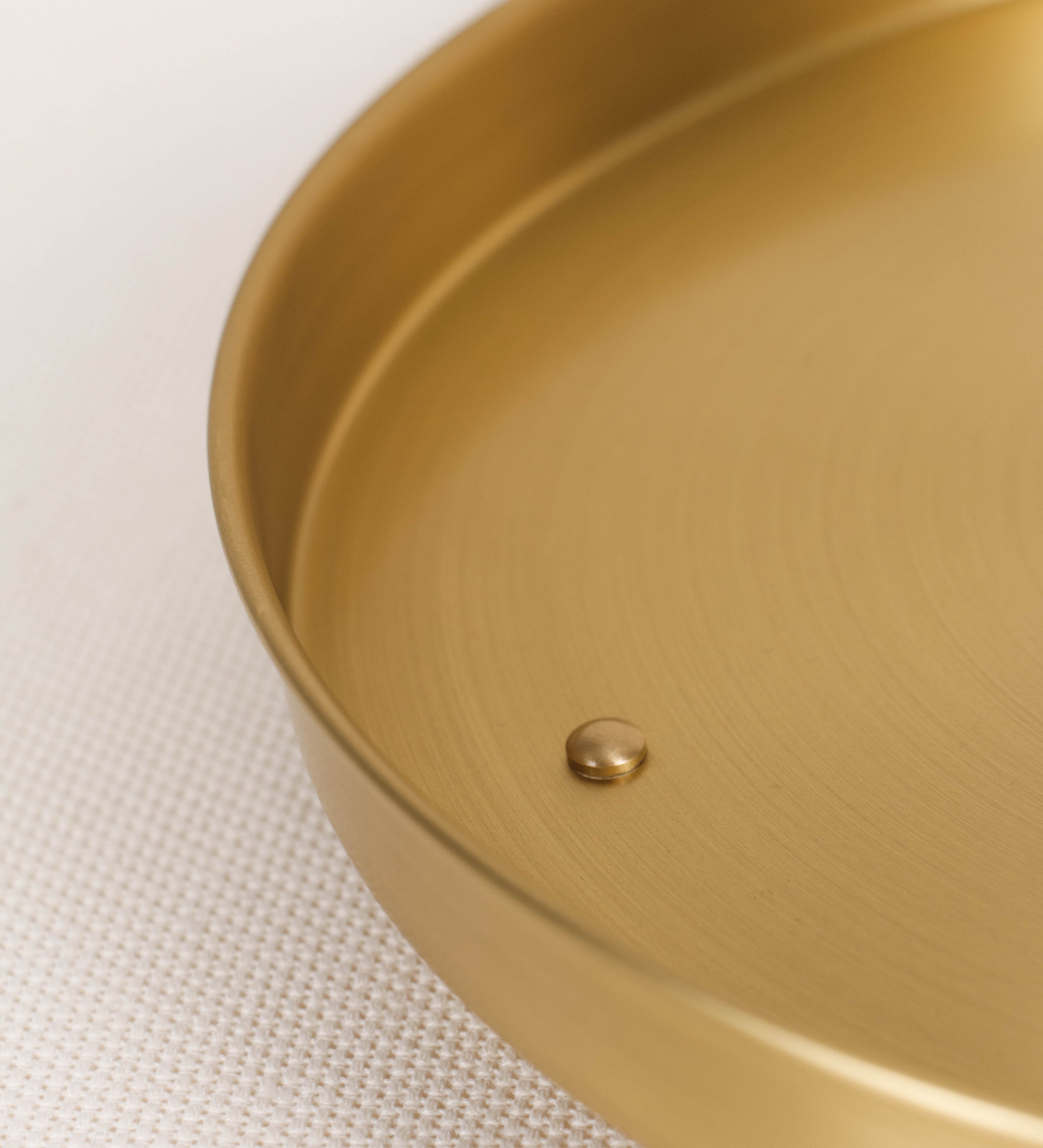Bandeja com um design sofisticado e intemporal. A bandeja é de latão, com um tom dourado mate e acabamento escovado. Tem uma forma circular, de superfície plana e com uma borda envolvente de altura média e grossura fina. Na sua base tem três pequenas esferas, em latão também, dispostas em formato triangular, que sustentam o prato circular no topo.