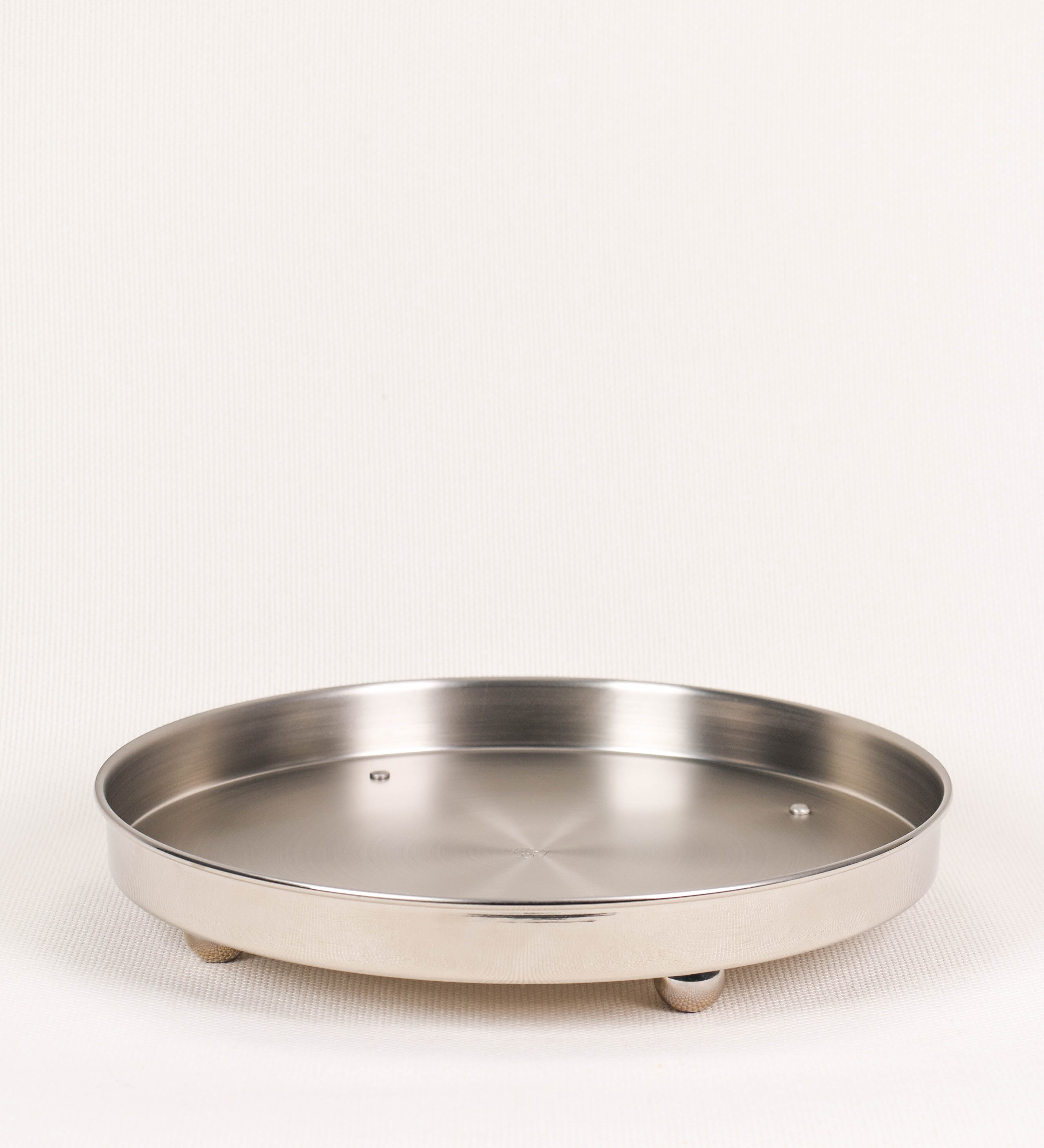 Bandeja com um design sofisticado e intemporal. A bandeja é de latão, tendo na superfície exterior um acabamento em tom prateado e com efeito de espelho, e um acabamento em tom prateado com efeito mate na superfície interior. Tem uma forma circular, de superfície plana e com uma borda envolvente de altura média e grossura fina. Na sua base tem três pequenas esferas, em níquel polido também, dispostas em formato triangular, que sustentam o prato circular no topo.