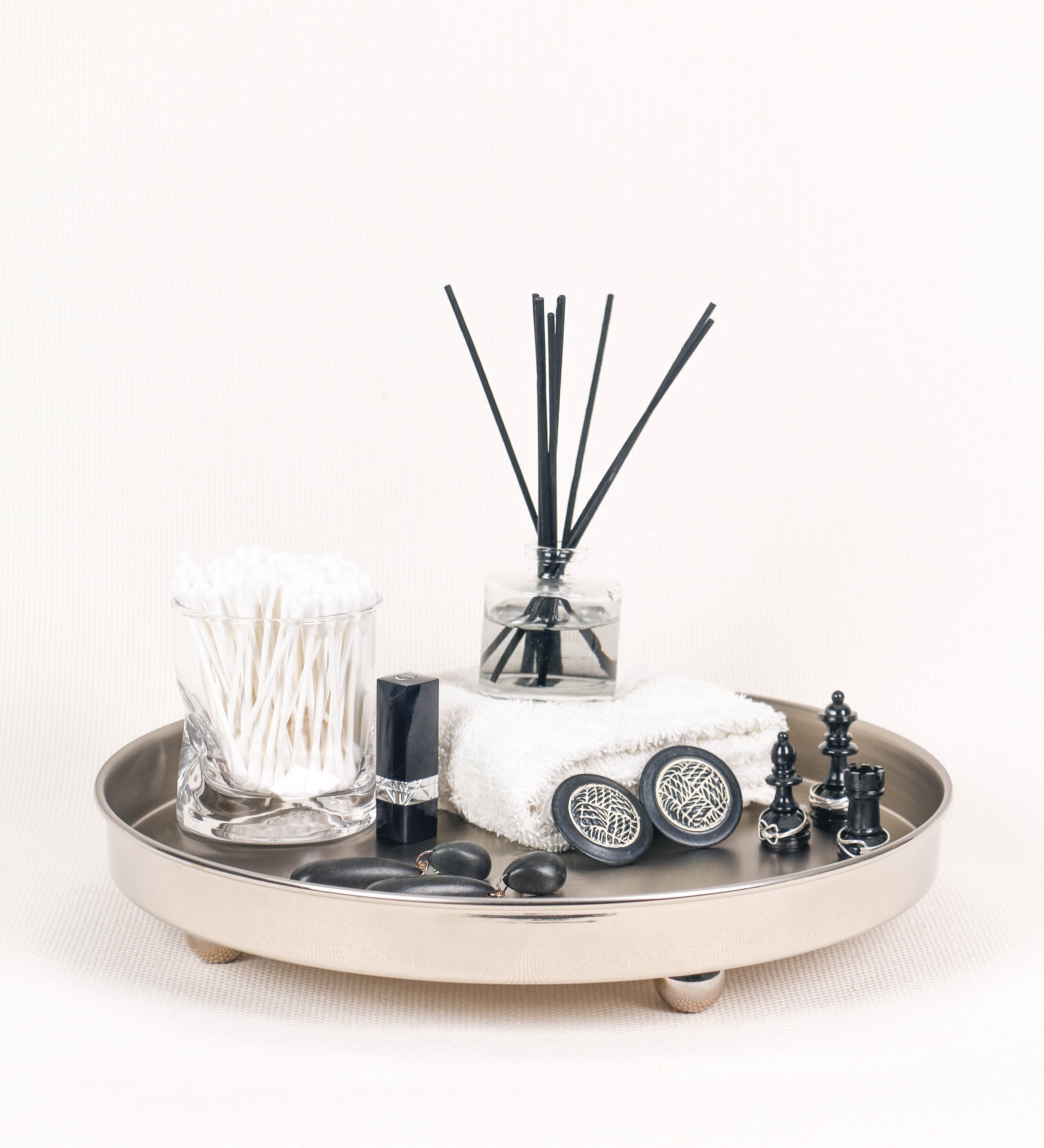 Bandeja com um design sofisticado e intemporal. A bandeja é de latão, tendo na superfície exterior um acabamento em tom prateado e com efeito de espelho, e um acabamento em tom prateado com efeito mate na superfície interior. Tem uma forma circular, de superfície plana e com uma borda envolvente de altura média e grossura fina. Na sua base tem três pequenas esferas, em níquel polido também, dispostas em formato triangular, que sustentam o prato circular no topo.