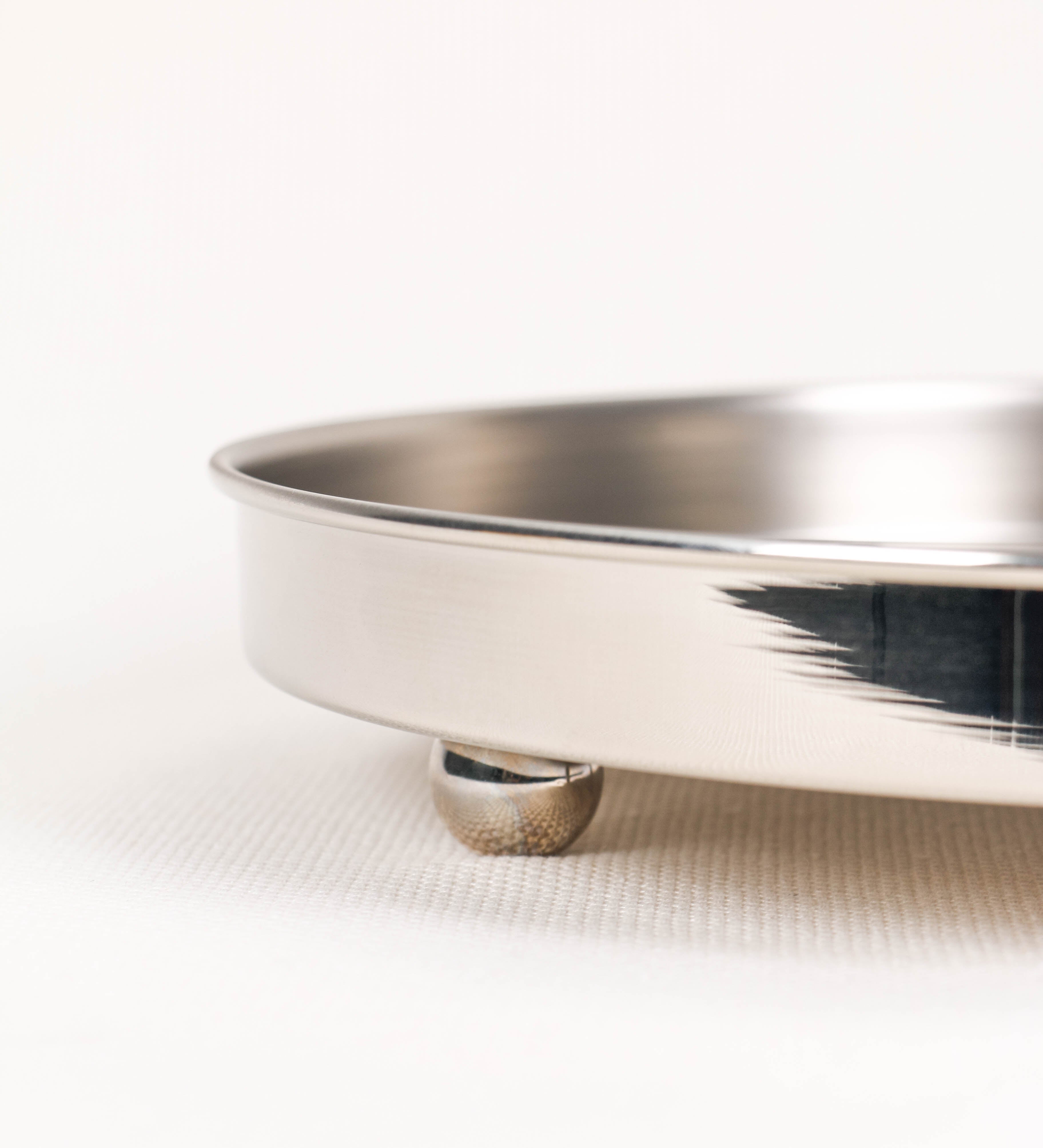 Bandeja com um design sofisticado e intemporal. A bandeja é de latão, tendo na superfície exterior um acabamento em tom prateado e com efeito de espelho, e um acabamento em tom prateado com efeito mate na superfície interior. Tem uma forma circular, de superfície plana e com uma borda envolvente de altura média e grossura fina. Na sua base tem três pequenas esferas, em níquel polido também, dispostas em formato triangular, que sustentam o prato circular no topo.