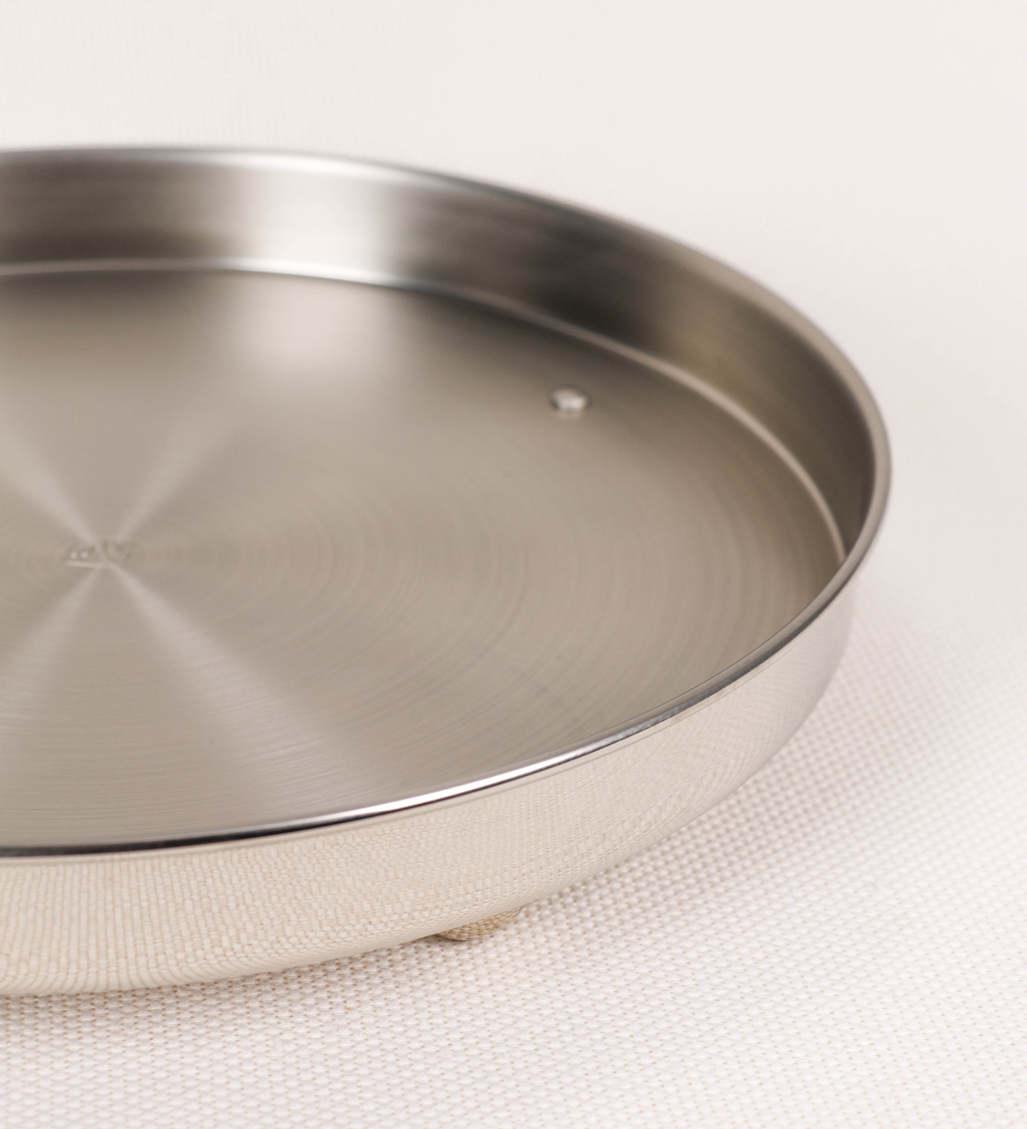 Bandeja com um design sofisticado e intemporal. A bandeja é de latão, tendo na superfície exterior um acabamento em tom prateado e com efeito de espelho, e um acabamento em tom prateado com efeito mate na superfície interior. Tem uma forma circular, de superfície plana e com uma borda envolvente de altura média e grossura fina. Na sua base tem três pequenas esferas, em níquel polido também, dispostas em formato triangular, que sustentam o prato circular no topo.