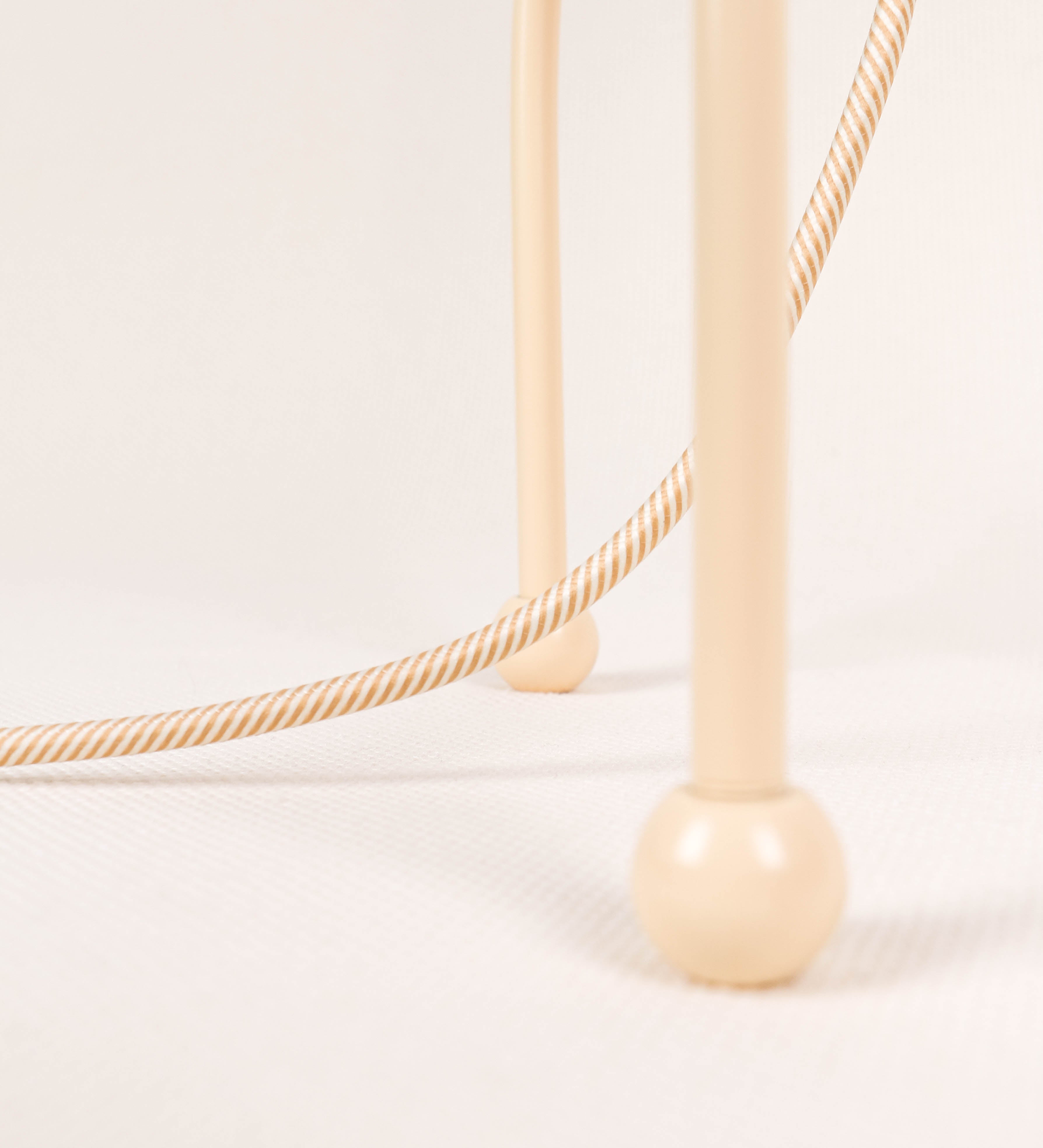 Candeeiro de mesa, de tom bege claro, com um design moderno e minimalista. O candeeiro tem o corpo com três pés curvados, terminando em pequenas esferas. A estrutura dos pés é interligada por uma peça central esférica. O cabo é revestido têxtil com um padrão de linhas douradas e brancas entrelaçadas, com interruptor e ficha transparentes.