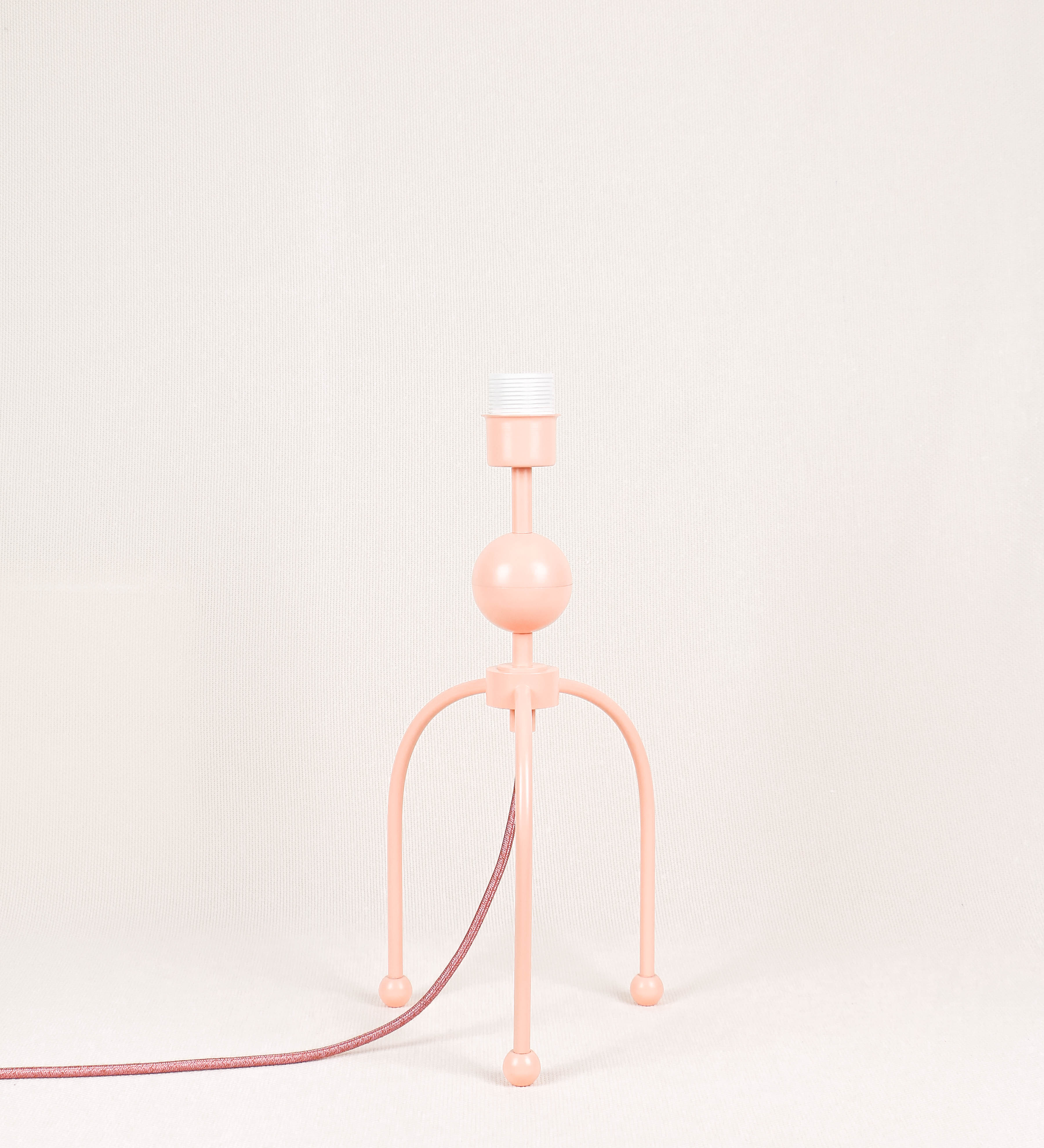 Candeeiro de mesa, de tom rosa salmão, com um design moderno e minimalista. O candeeiro tem o corpo com três pés curvados, terminando em pequenas esferas. A estrutura dos pés é interligada por uma peça central esférica. O cabo é revestido têxtil de tom rosa velho, com interruptor e ficha transparentes.