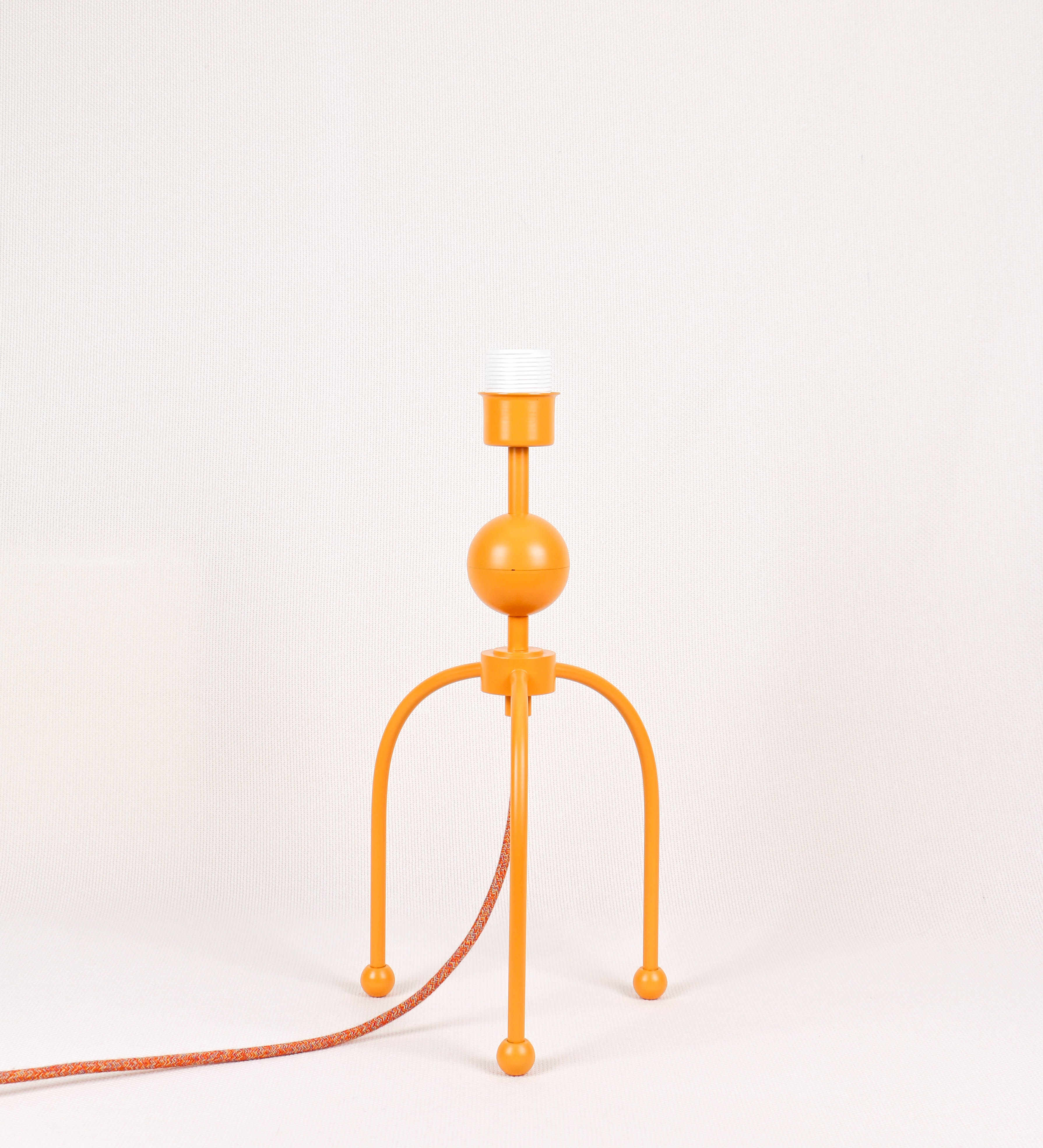 Candeeiro de mesa, de tom laranja-tangerina, com um design moderno e minimalista. O candeeiro tem o corpo com três pés curvados, terminando em pequenas esferas. A estrutura dos pés é interligada por uma peça central esférica. O cabo é revestido têxtil com um padrão de linhas coloridas (laranjas, azuis, amarelas e outras), com interruptor e ficha transparentes.