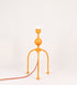 Candeeiro de mesa, de tom laranja-tangerina, com um design moderno e minimalista. O candeeiro tem o corpo com três pés curvados, terminando em pequenas esferas. A estrutura dos pés é interligada por uma peça central esférica. O cabo é revestido têxtil com um padrão de linhas coloridas (laranjas, azuis, amarelas e outras), com interruptor e ficha transparentes.