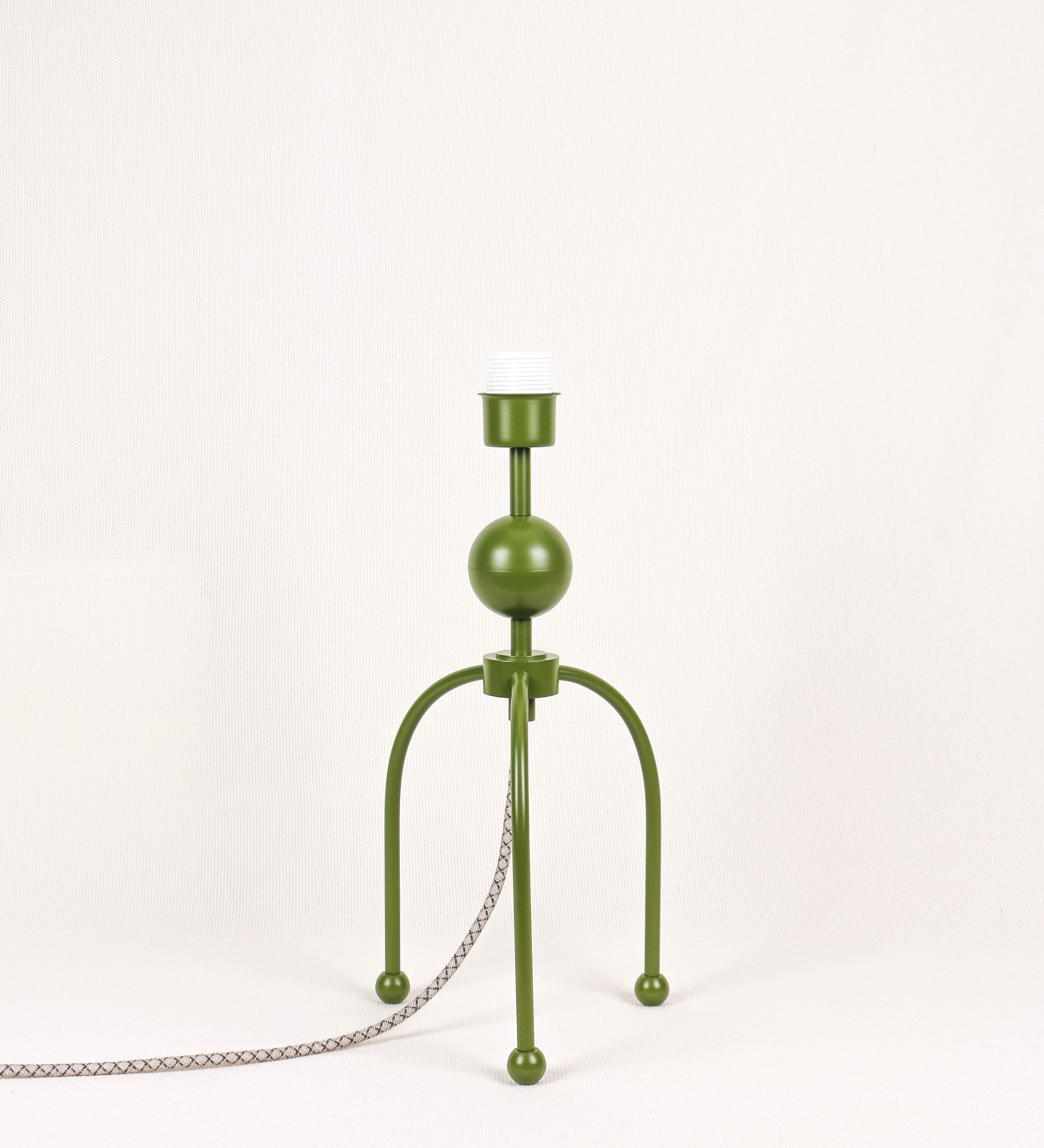 Candeeiro de mesa, de tom verde couve-de-bruxelas, com um design moderno e minimalista. O candeeiro tem o corpo com três pés curvados, terminando em pequenas esferas. A estrutura dos pés é interligada por uma peça central esférica. O cabo é revestido têxtil com um padrão de linhas castanhas e beges entrelaçadas, com interruptor e ficha transparentes.