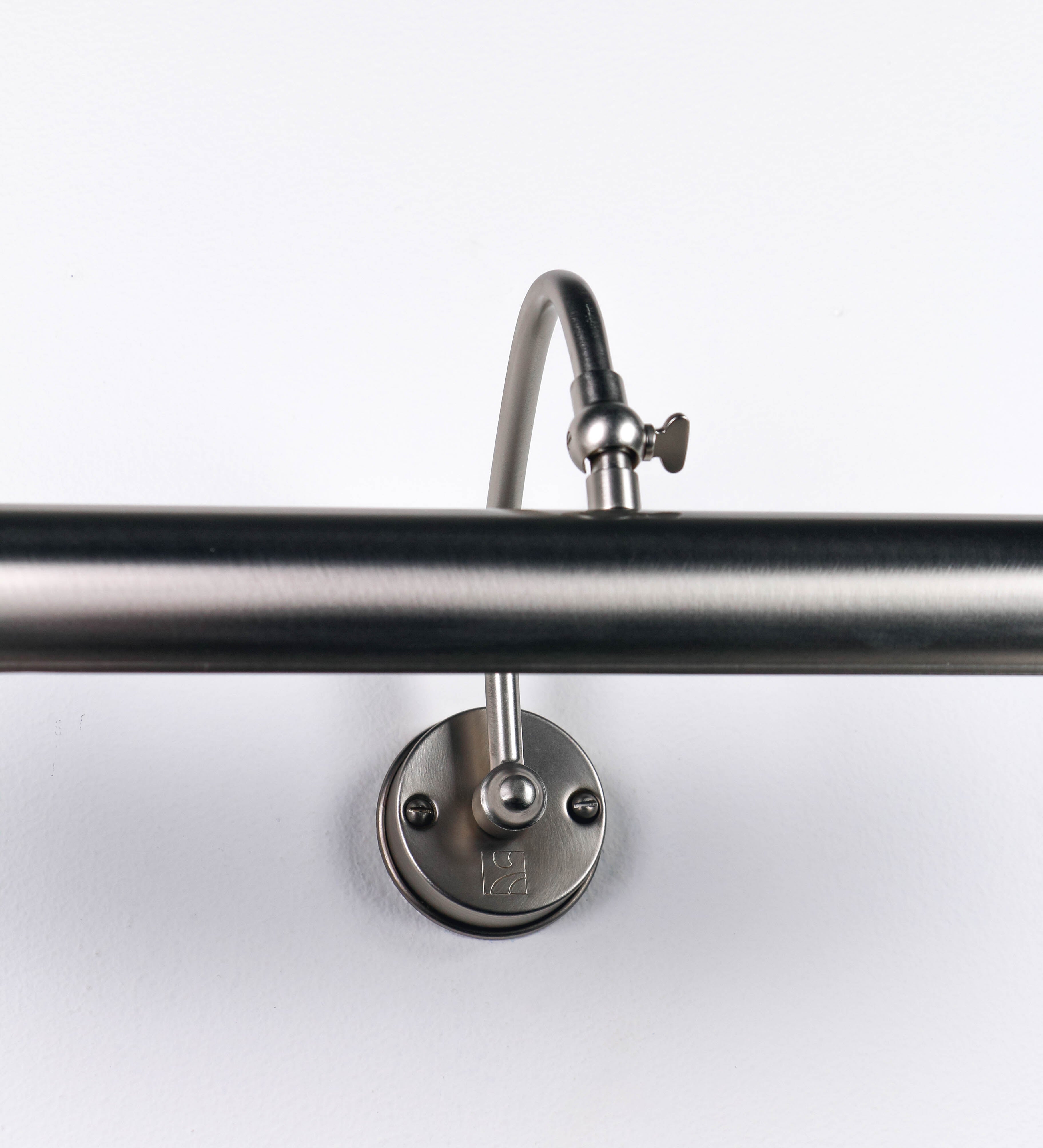 Aplique de parede que combina elementos clássicos com toques contemporâneos, todo em níquel escovado. O aplique é composto por uma base circular de onde se estende um braço curvo e gracioso, que suporta a barra principal. O braço contém um elemento que permite apertar ou desapertar a barra para melhor manipular a direção da luz. A barra de 45cm de largura é cilíndrica e alongada. As extremidades do cilindro são planas e do mesmo diâmetro.
