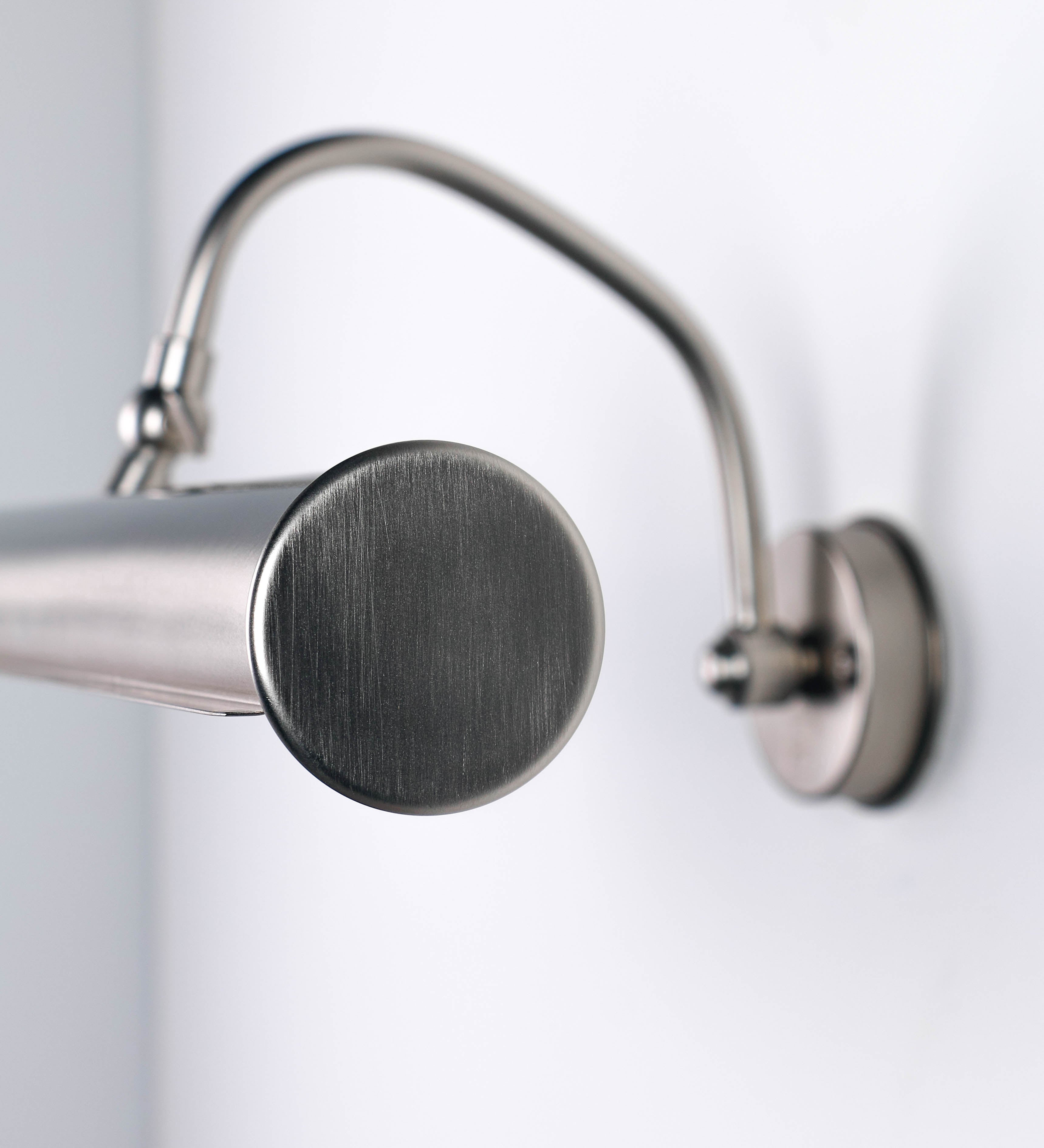 Aplique de parede que combina elementos clássicos com toques contemporâneos, todo em níquel escovado. O aplique é composto por uma base circular de onde se estende um braço curvo e gracioso, que suporta a barra principal. O braço contém um elemento que permite apertar ou desapertar a barra para melhor manipular a direção da luz. A barra de 45cm de largura é cilíndrica e alongada. As extremidades do cilindro são planas e do mesmo diâmetro.
