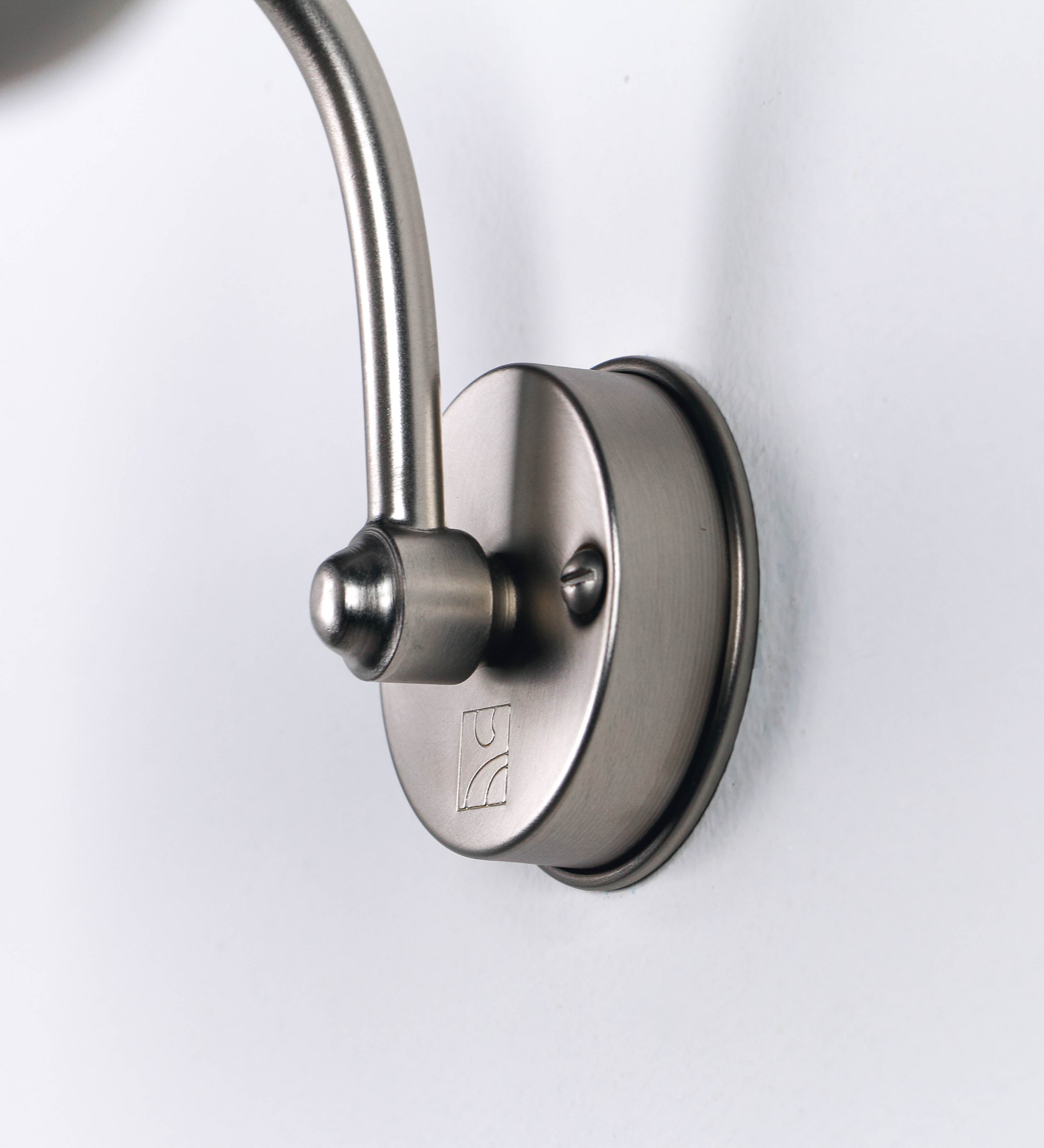 Aplique de parede que combina elementos clássicos com toques contemporâneos, todo em níquel escovado. O aplique é composto por uma base circular de onde se estende um braço curvo e gracioso, que suporta a barra principal. O braço contém um elemento que permite apertar ou desapertar a barra para melhor manipular a direção da luz. A barra de 45cm de largura é cilíndrica e alongada. As extremidades do cilindro são planas e do mesmo diâmetro.