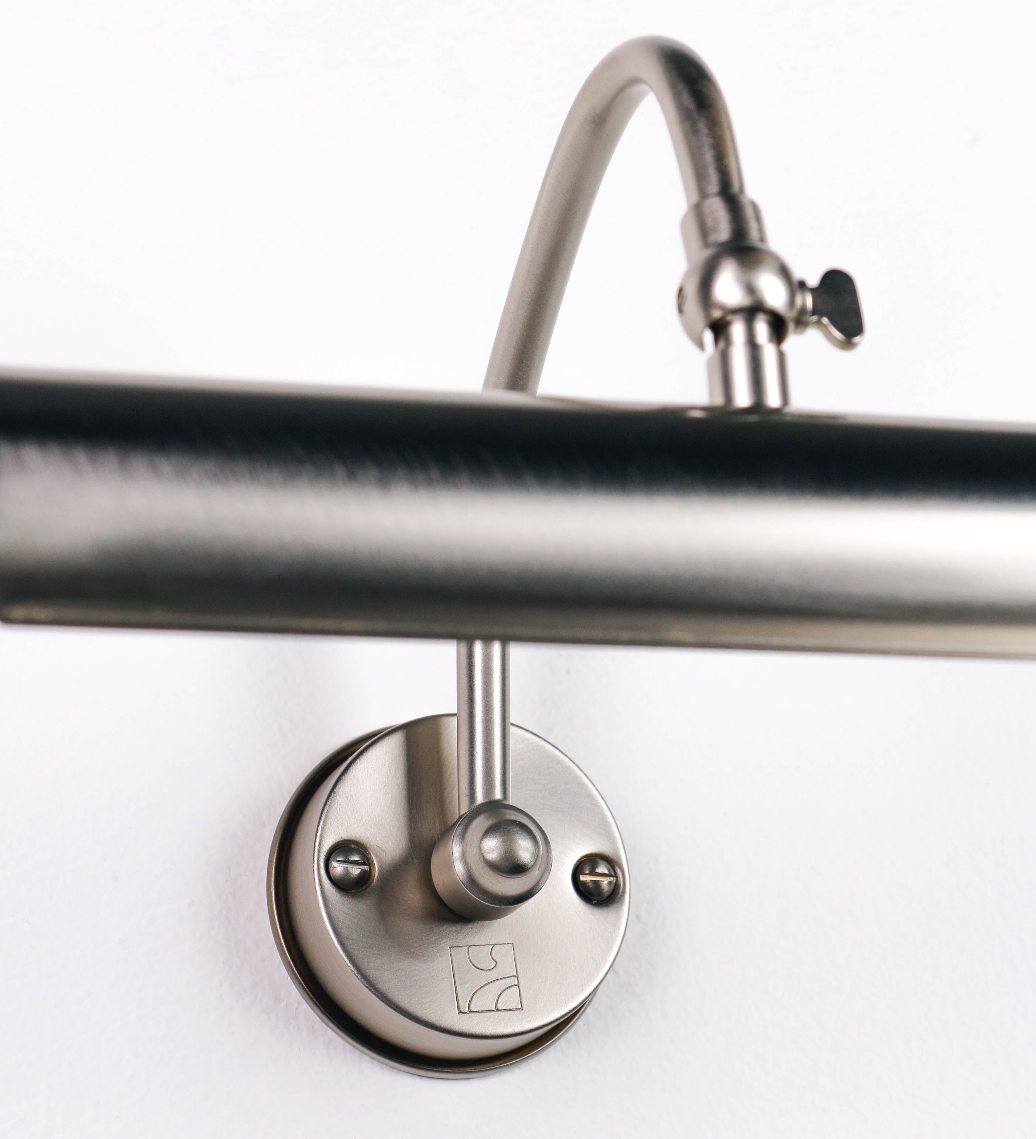 Aplique de parede que combina elementos clássicos com toques contemporâneos, todo em níquel escovado. O aplique é composto por uma base circular de onde se estende um braço curvo e gracioso, que suporta a barra principal. O braço contém um elemento que permite apertar ou desapertar a barra para melhor manipular a direção da luz. A barra de 25cm de largura é cilíndrica e alongada. As extremidades do cilindro são planas e do mesmo diâmetro.